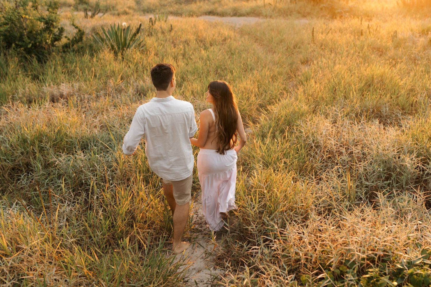 A couple stands together in a golden field at sunset while holding hands, photographed from behind.
