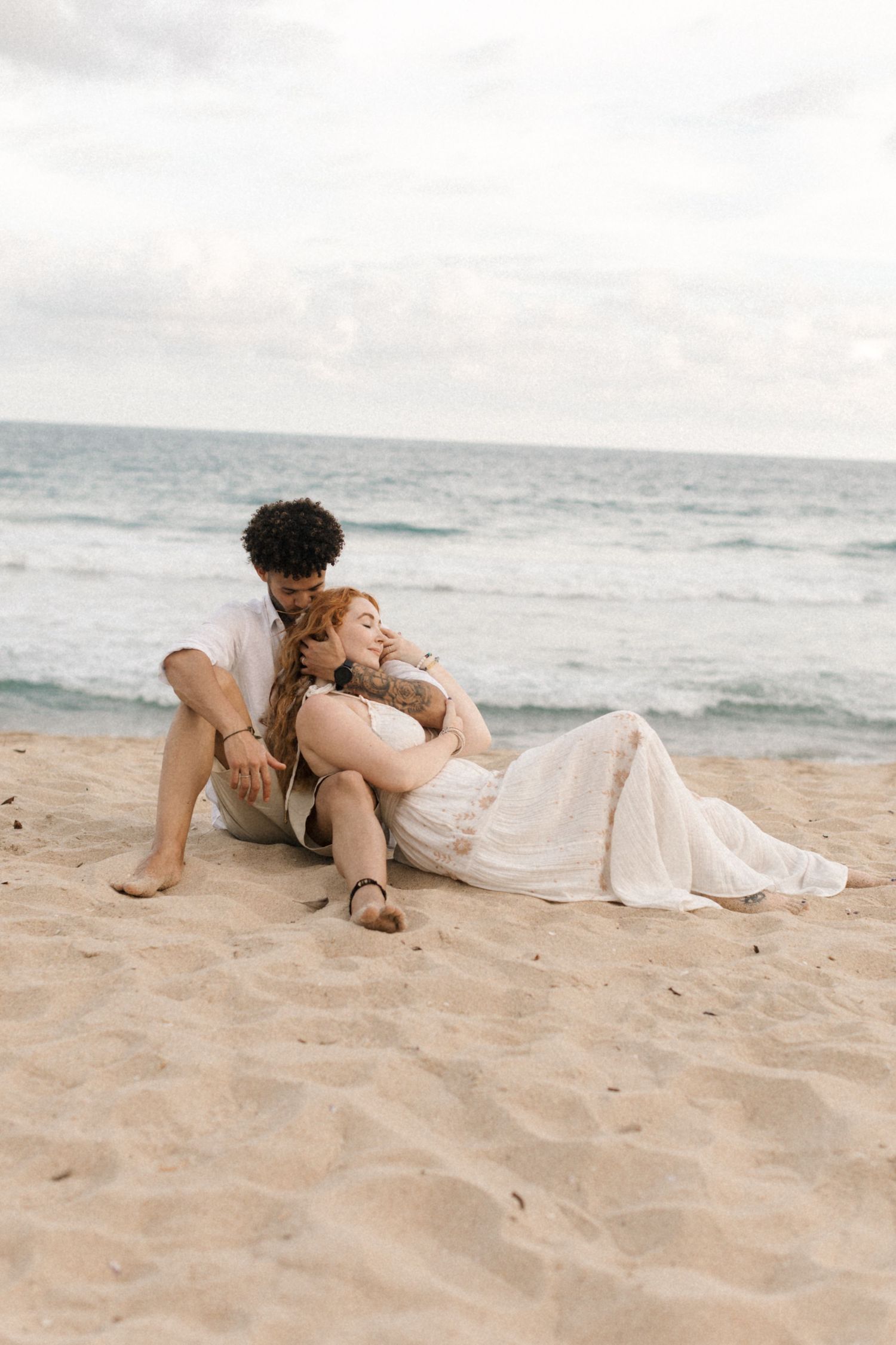 A couple relaxes together on the sandy beach with ocean waves in the background.
