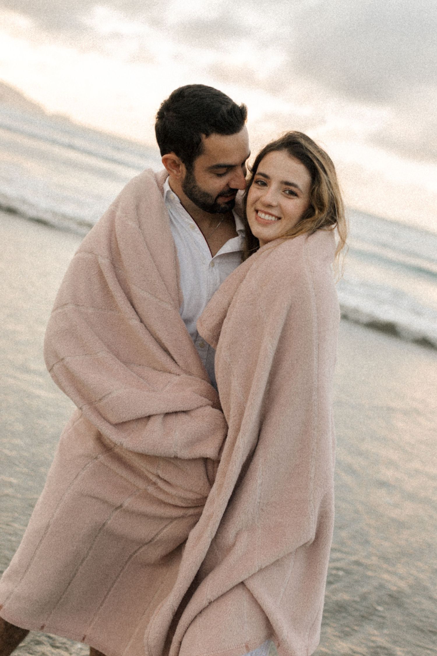 A couple shares a tender moment wrapped in a cozy pink blanket on a beach during golden hour.