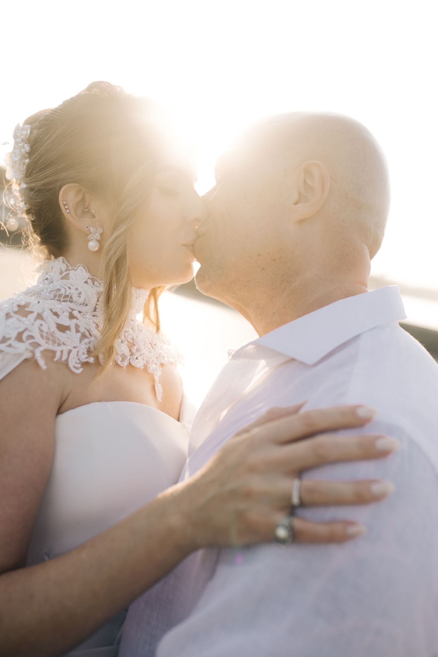 A backlit romantic kiss between newlyweds during golden hour by the water.