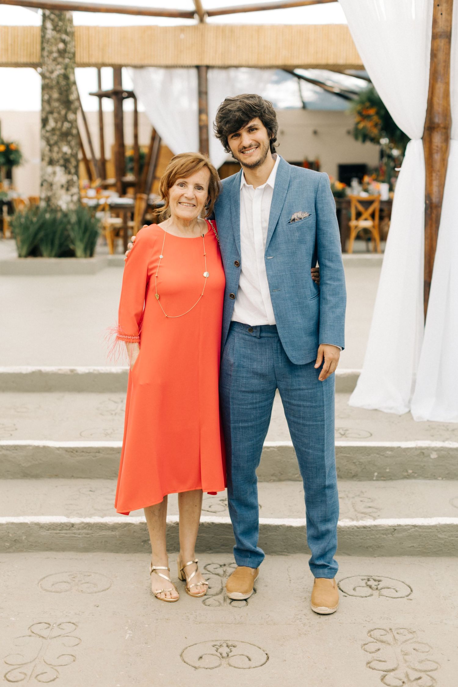 A pair standing together at an outdoor venue with white curtains and wooden chairs.