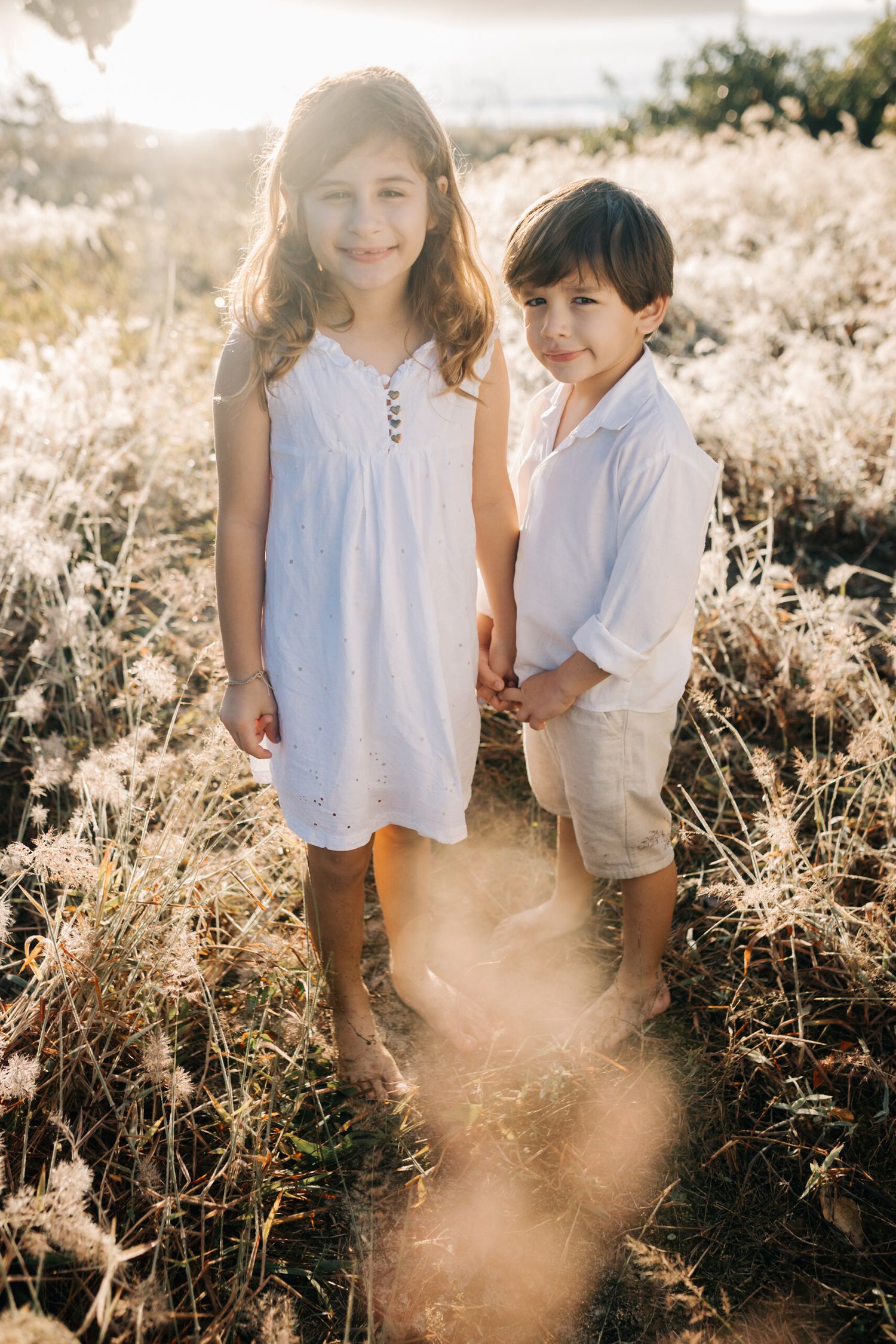 Two children hold hands while standing in tall grass during golden hour.