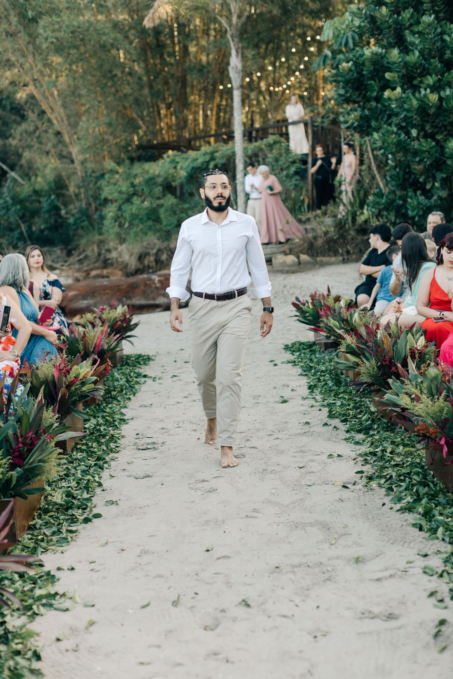 Wedding party members walking down sandy aisle with tropical plants and guests seated on sides.