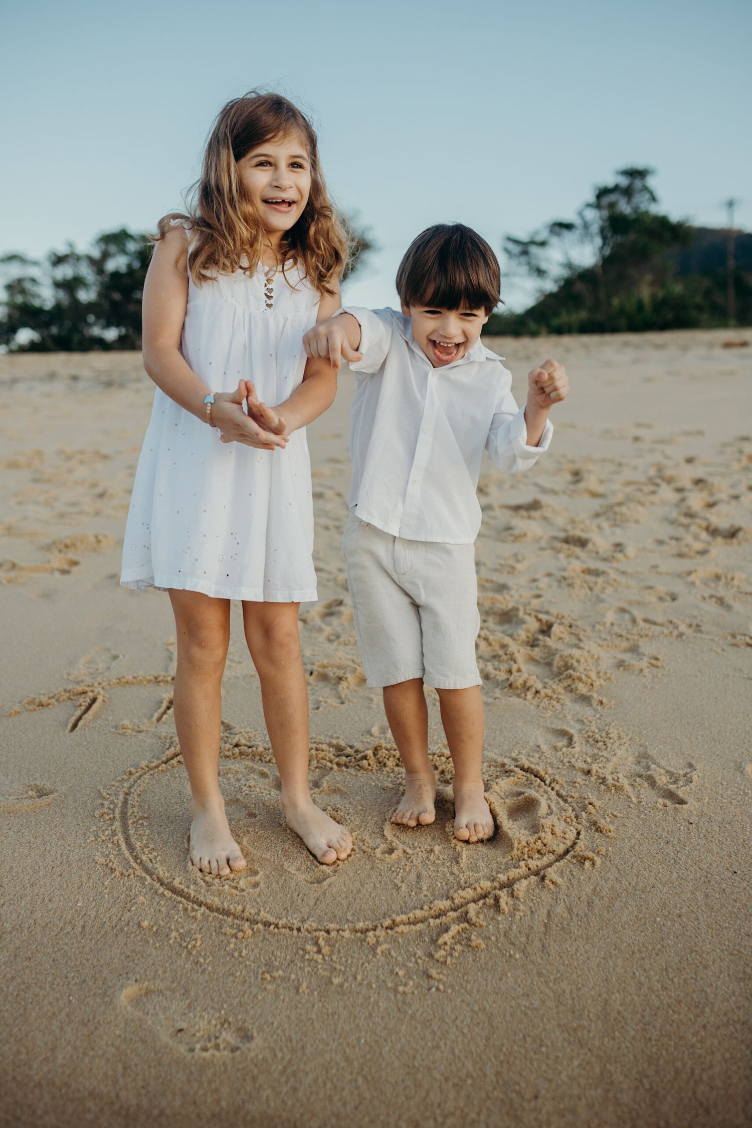 Siblings in matching white outfits play around a heart drawn in beach sand.