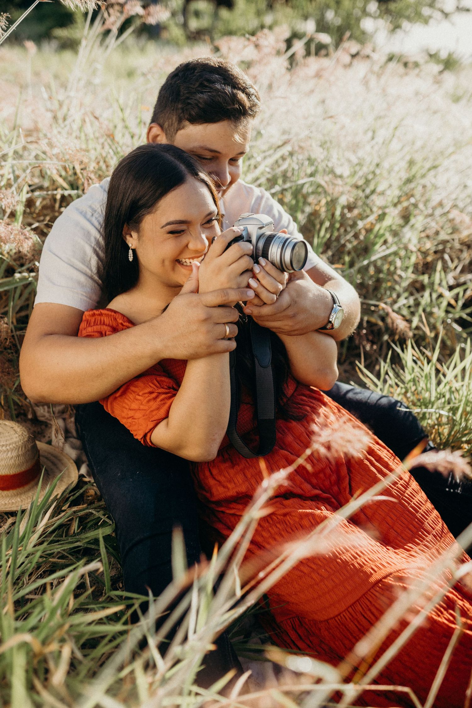 A joyful couple laughs together while sharing a camera in a sunlit meadow.