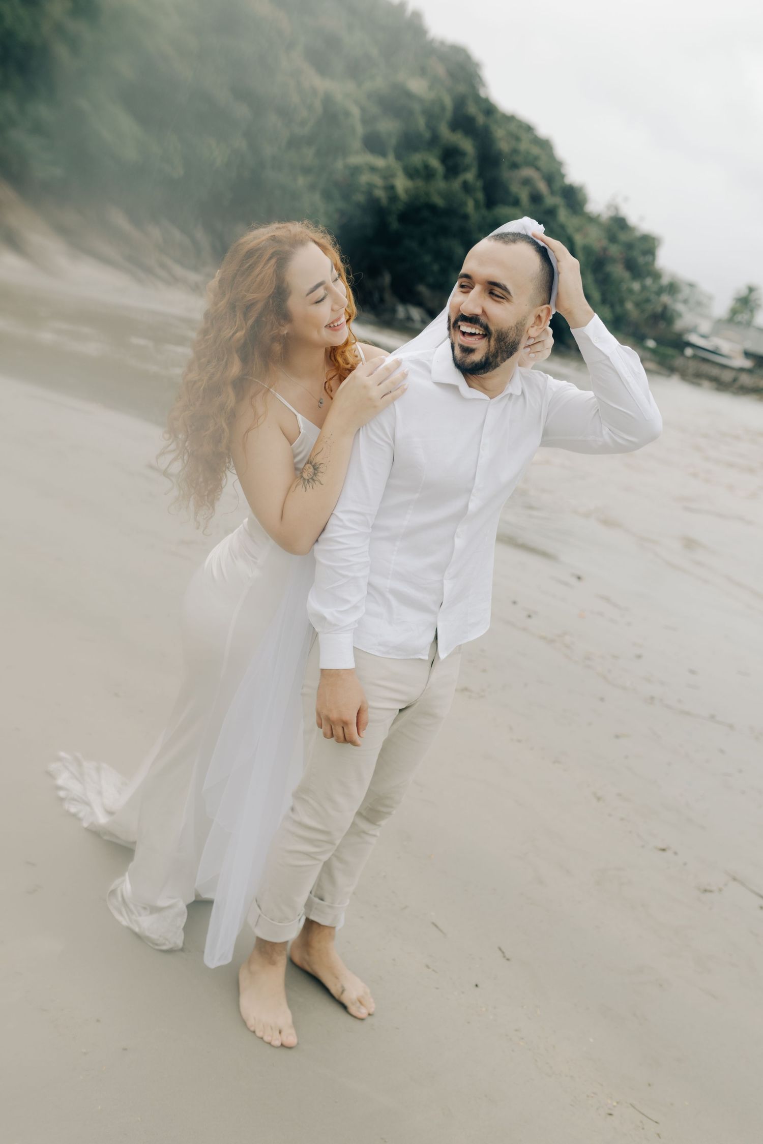 A romantic couple in white attire dances and laughs together on an overcast beach.
