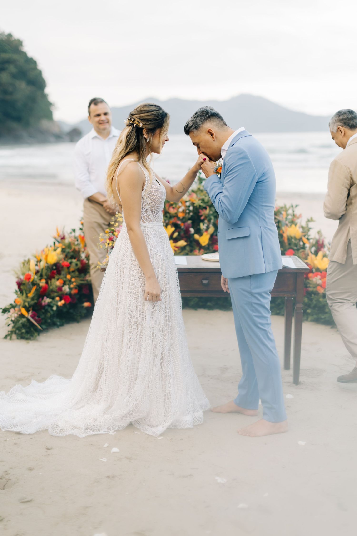 Candid beach wedding ceremony with tropical floral decorations and mountains in background.