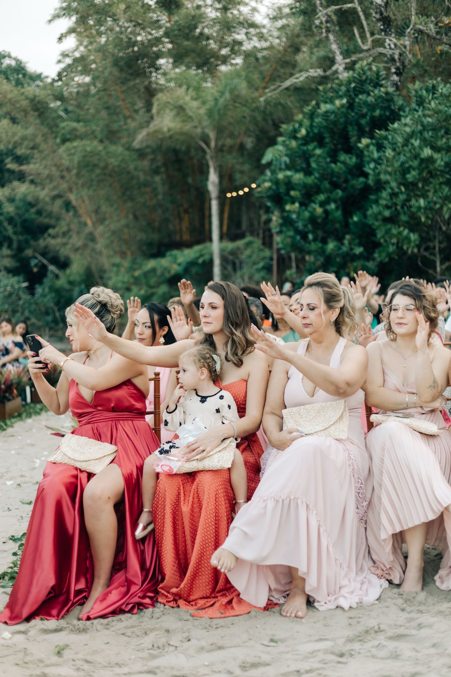 Wedding guests in coral and blush dresses sitting in wooden chairs during an outdoor ceremony.