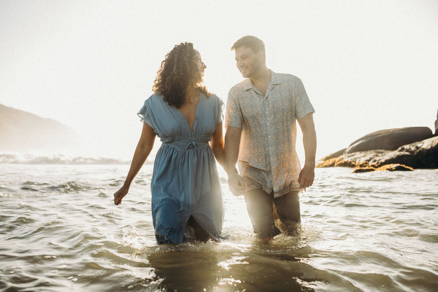 Happy couple walking through shallow ocean waters during sunset with mountains behind them.