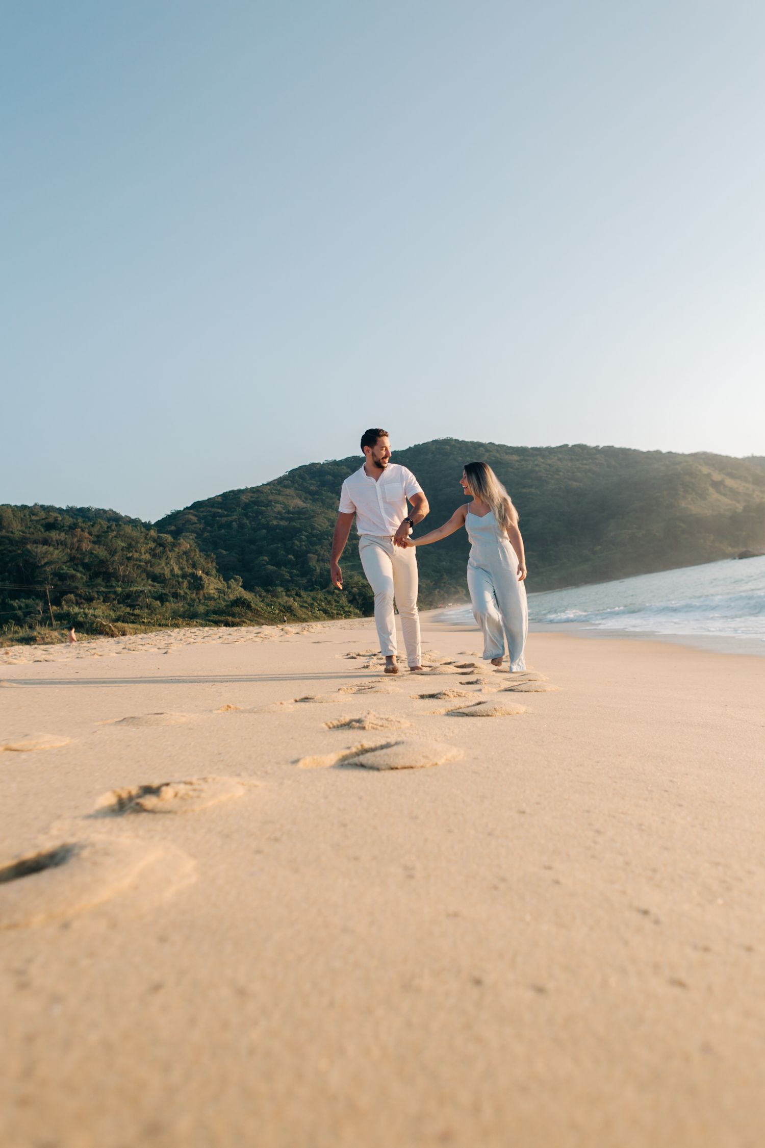 Two people strolling together on a sandy beach with coastal hills during golden hour.