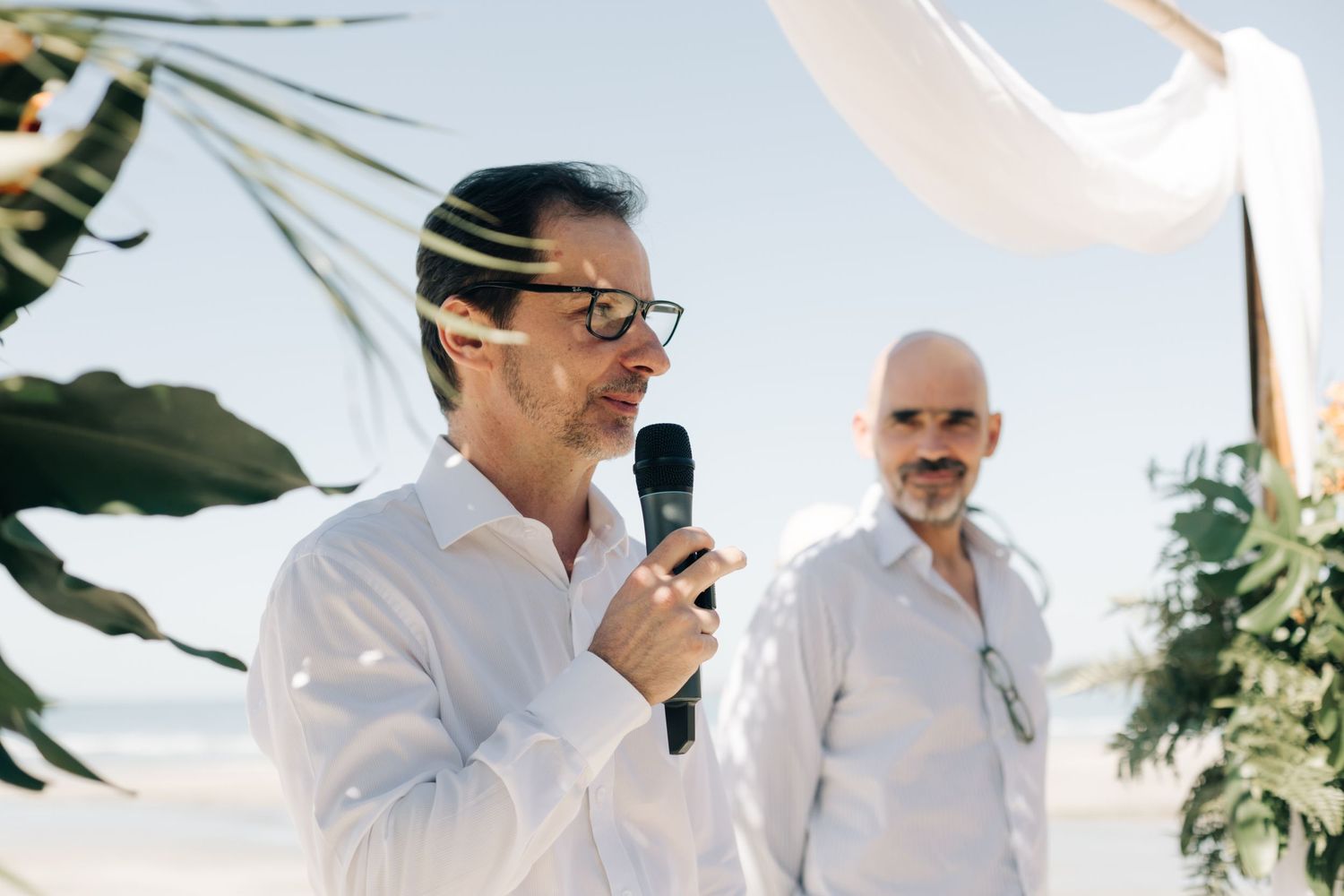 Two people in white shirts speaking at an outdoor wedding ceremony with tropical foliage in the background.