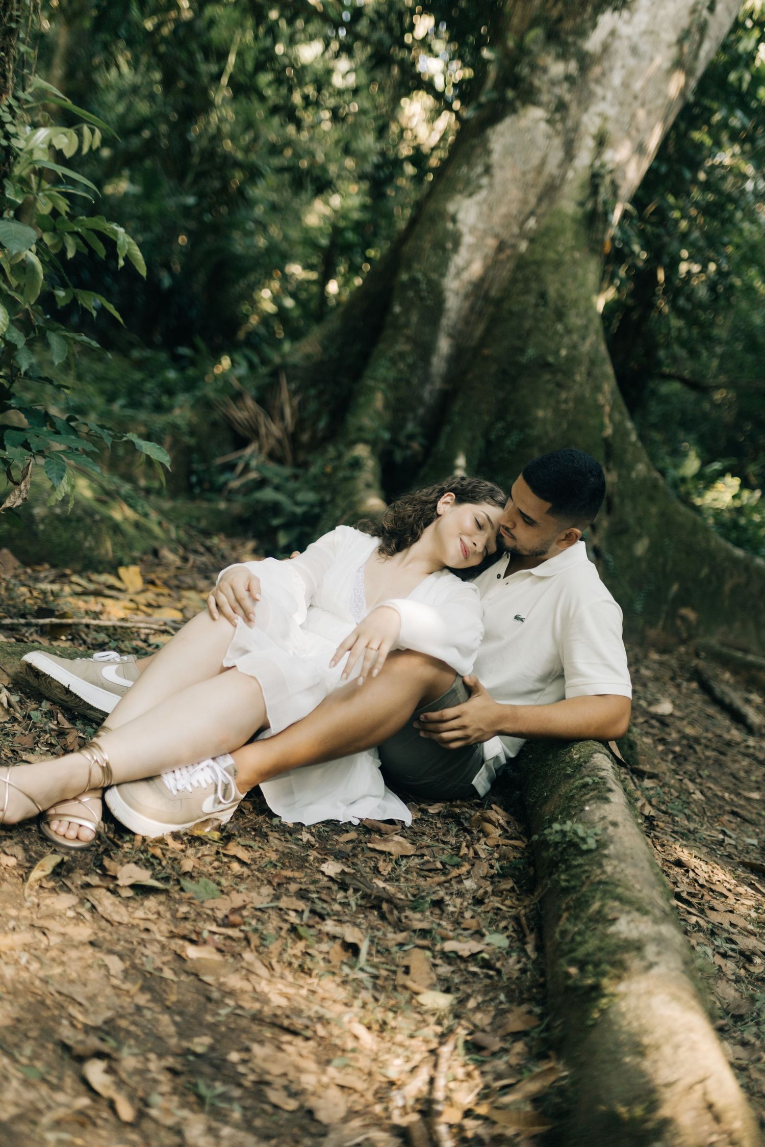 A couple relaxes together on a forest floor near a large tree trunk.