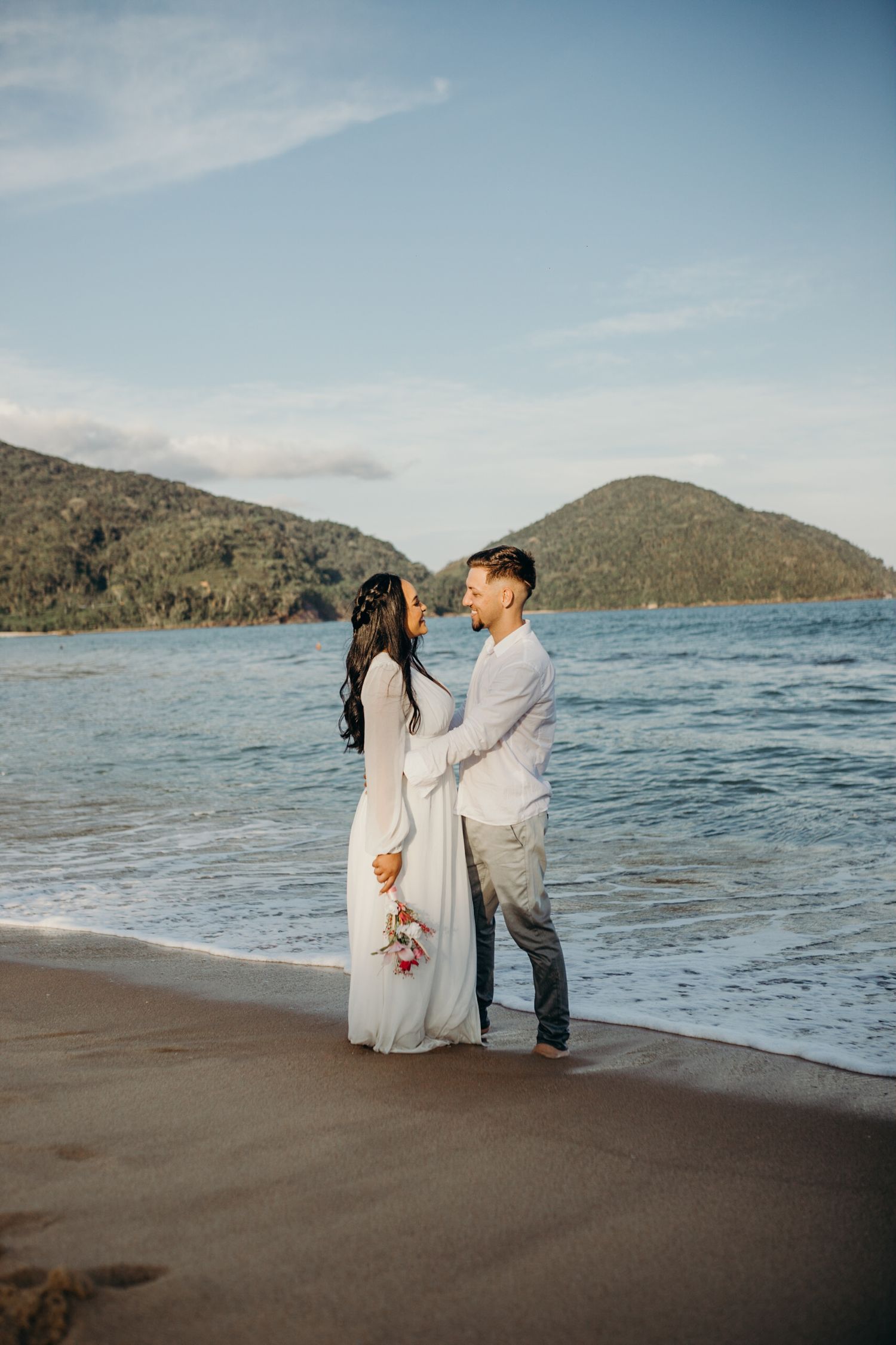 Couple in white clothing shares romantic moments on beach with mountains and ocean in background.