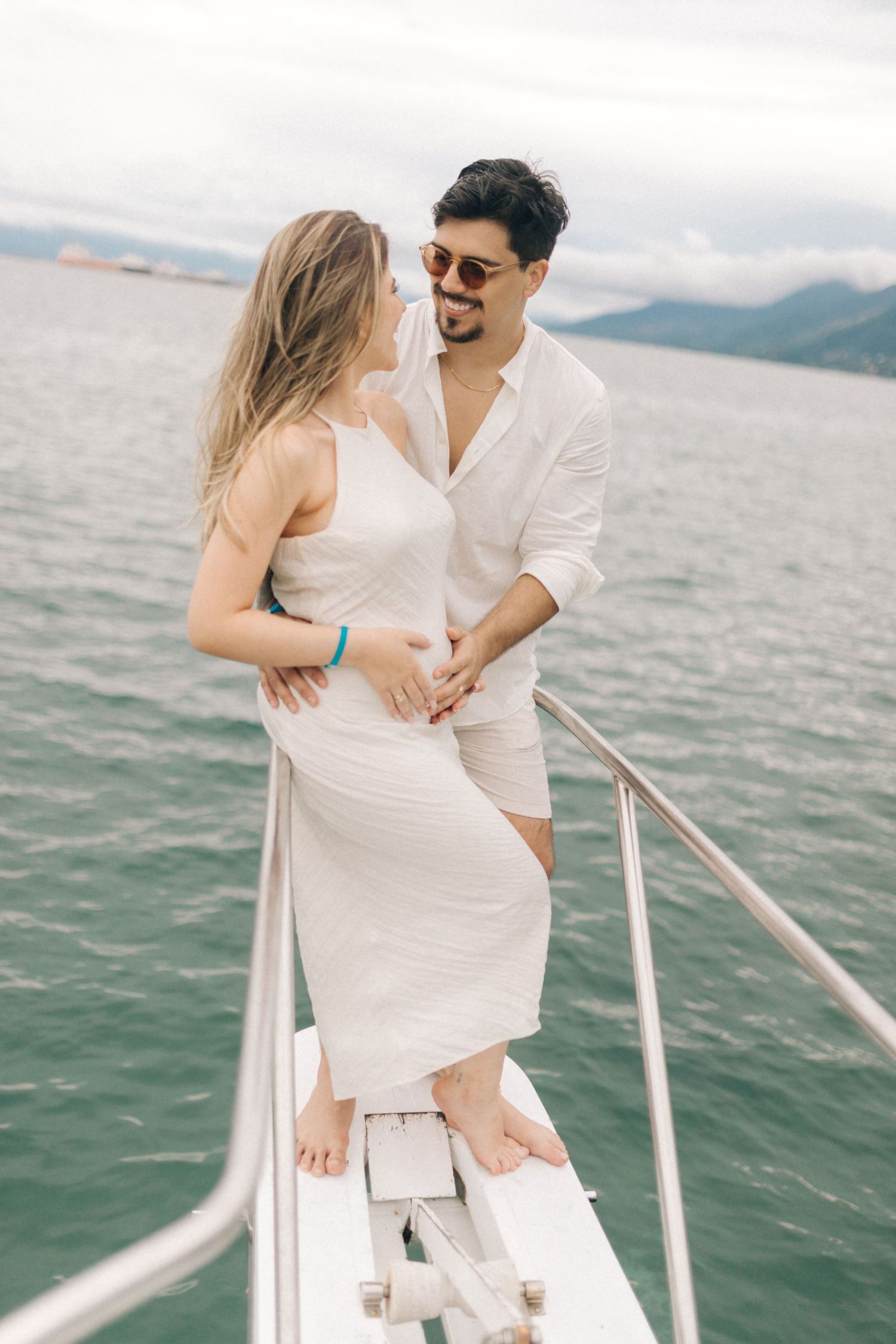 Romantic couple standing on yacht bow with scenic mountain and ocean backdrop.