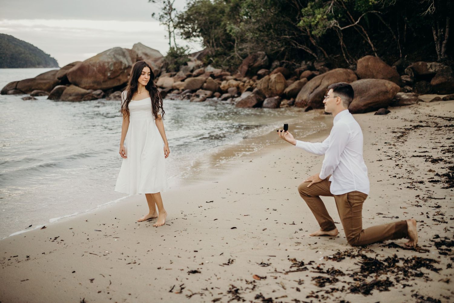 Couple stands together on beach with rocky shoreline and trees.