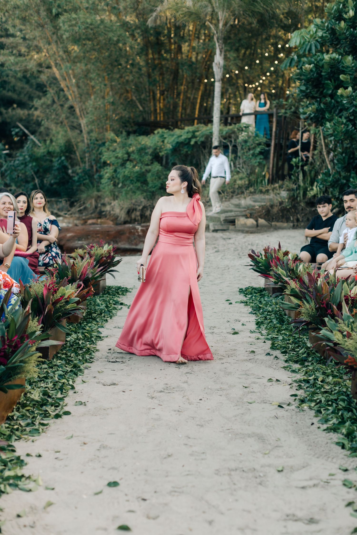 Person wearing coral gown proceeds down sand pathway lined with potted plants.