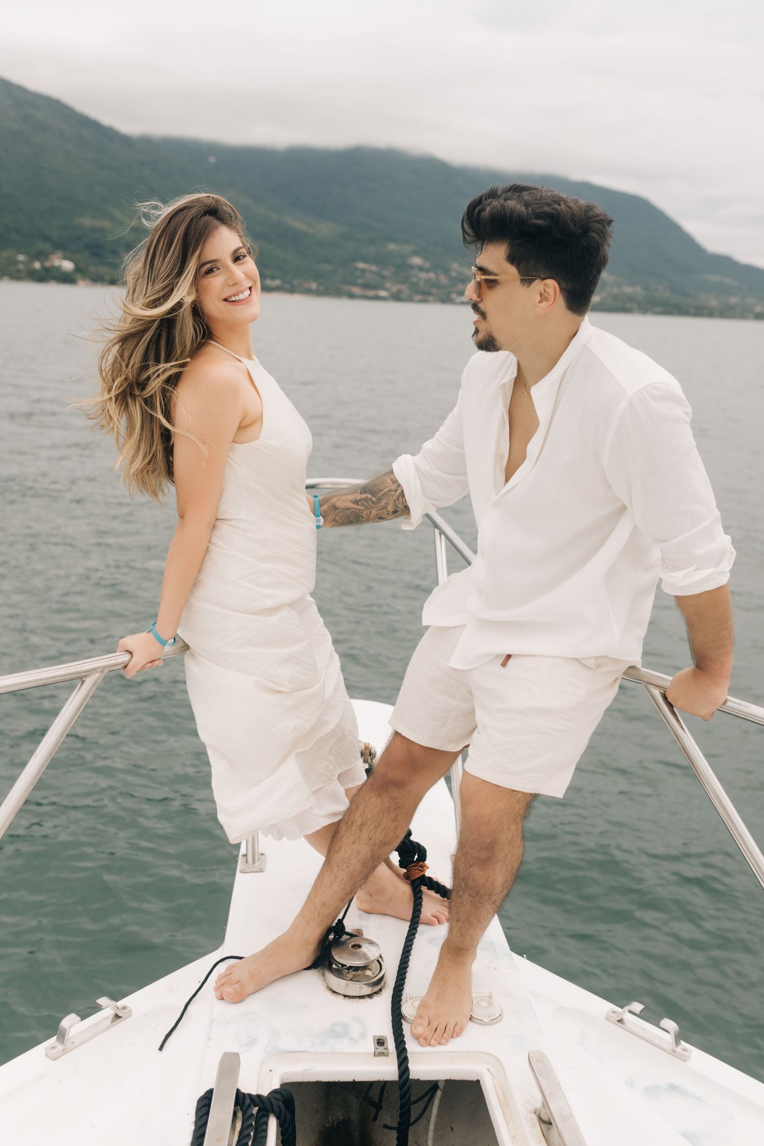 A couple dressed in white laugh together on the front of a boat.