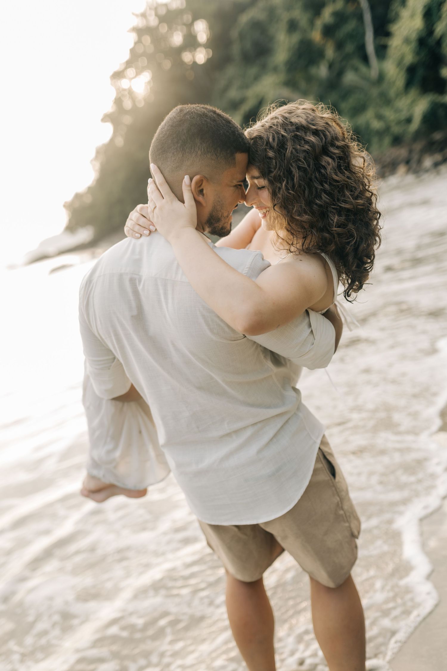 A romantic couple shares intimate dance poses on a sunlit beach near the water.