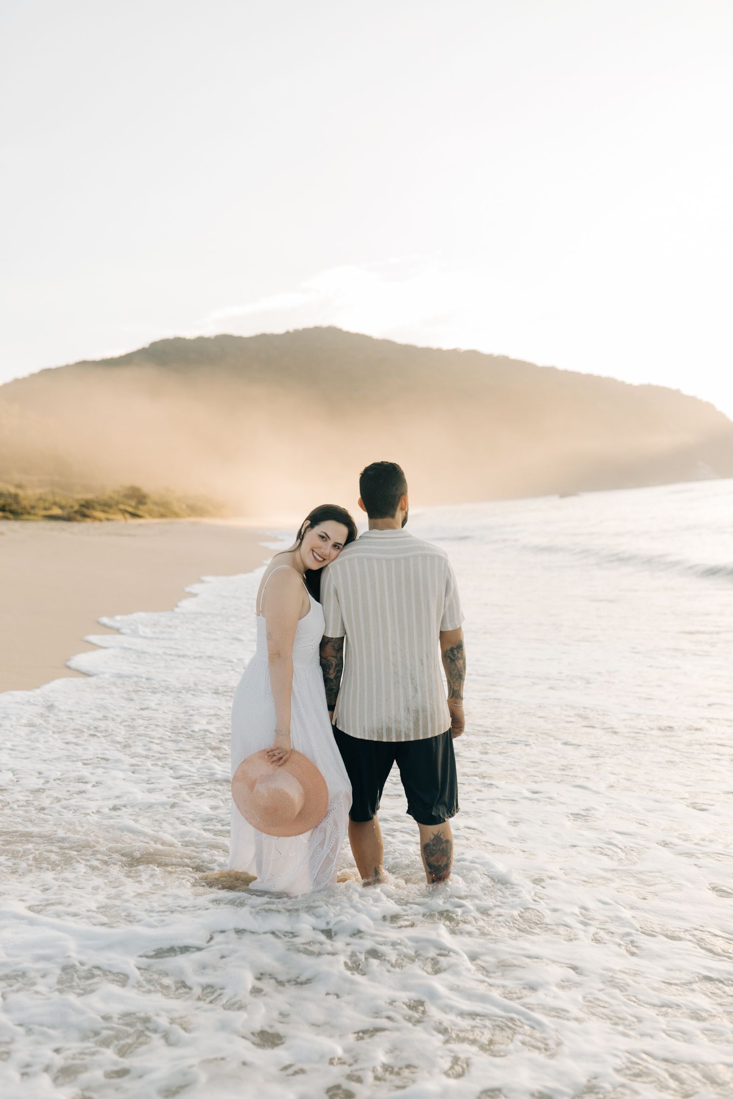 A couple sits together on a sandy beach during sunset with mountains in the background.