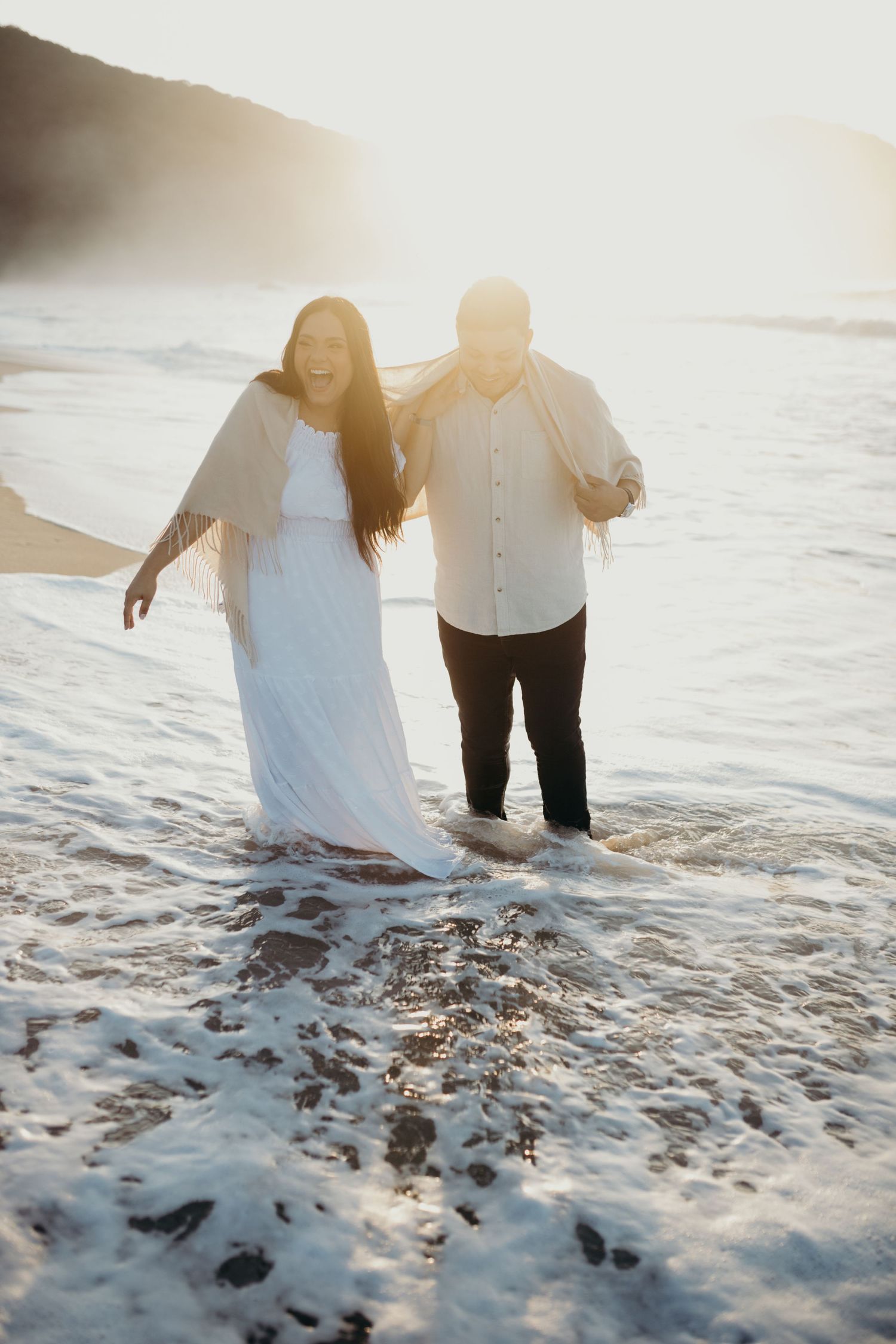 Couple walking through ocean waves at sunset wearing white and black clothing.