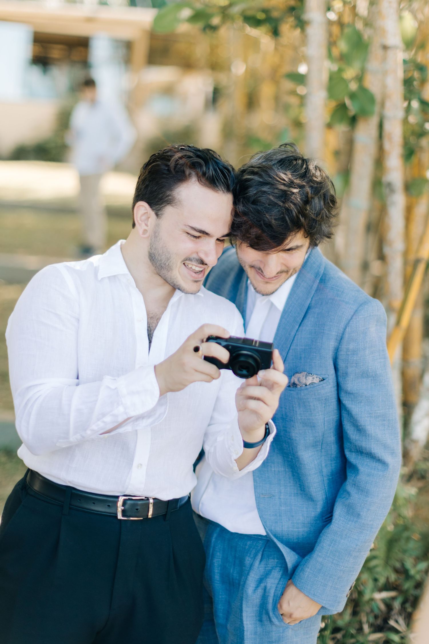 Two people in formal attire share a laugh while looking at something together outdoors.