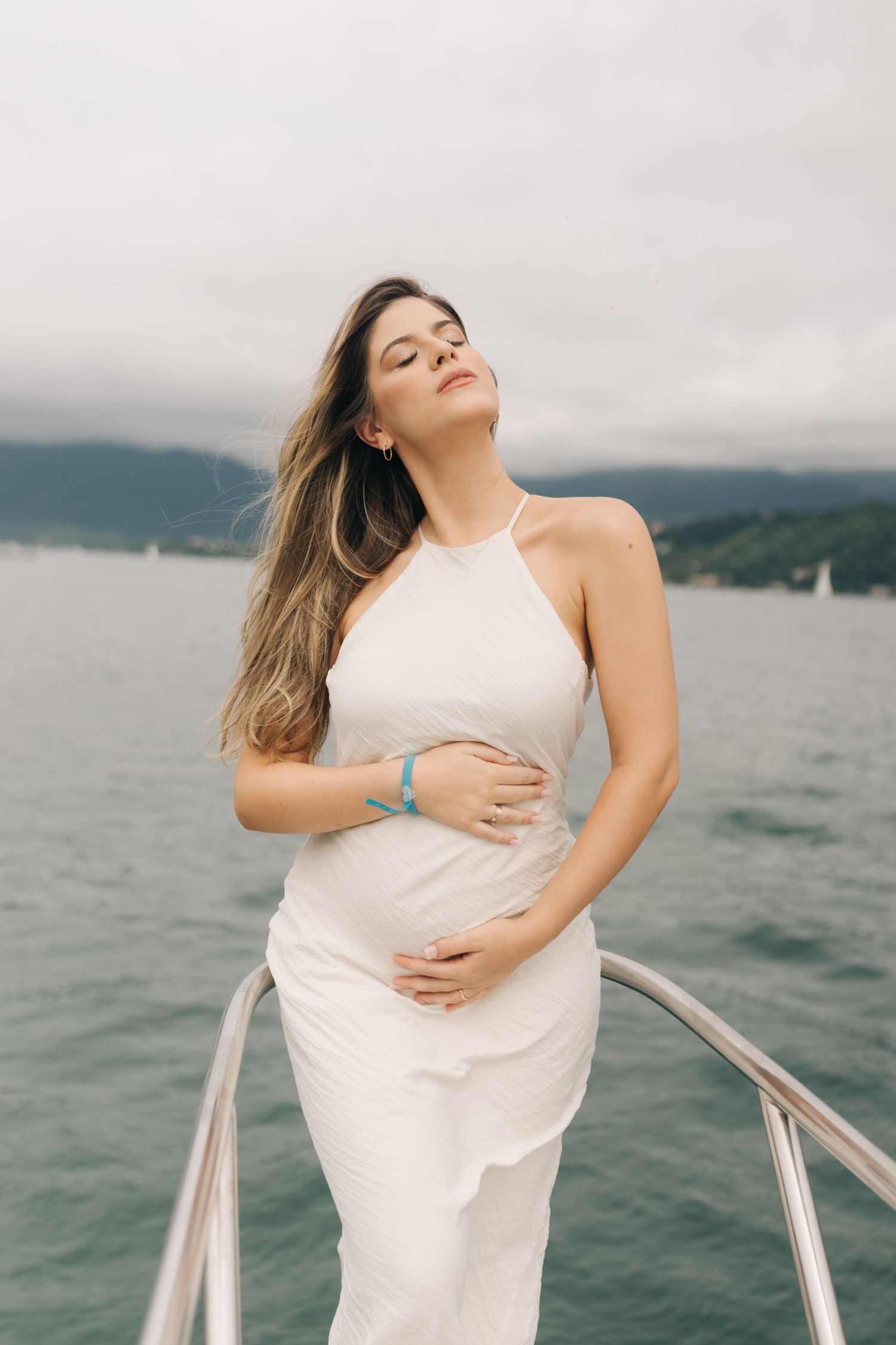 A woman in a flowing white dress poses on a boat deck with mountains and water in the background.