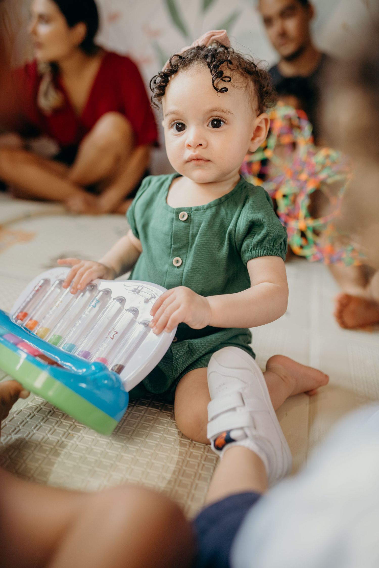 A young child in a green outfit examining a colorful toy while sitting on a floor mat.