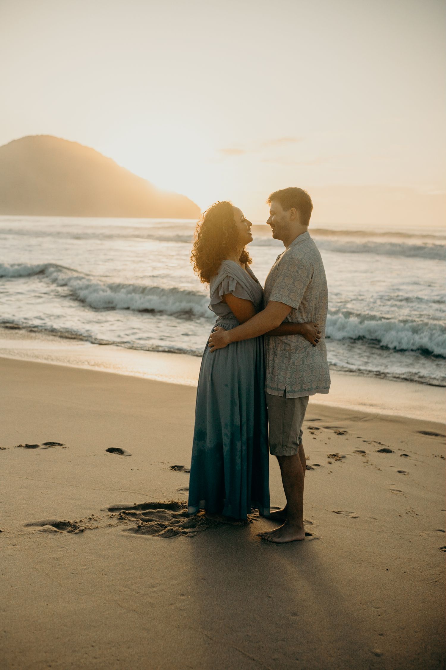 Silhouetted couple embracing on beach as sun sets behind coastal mountain.