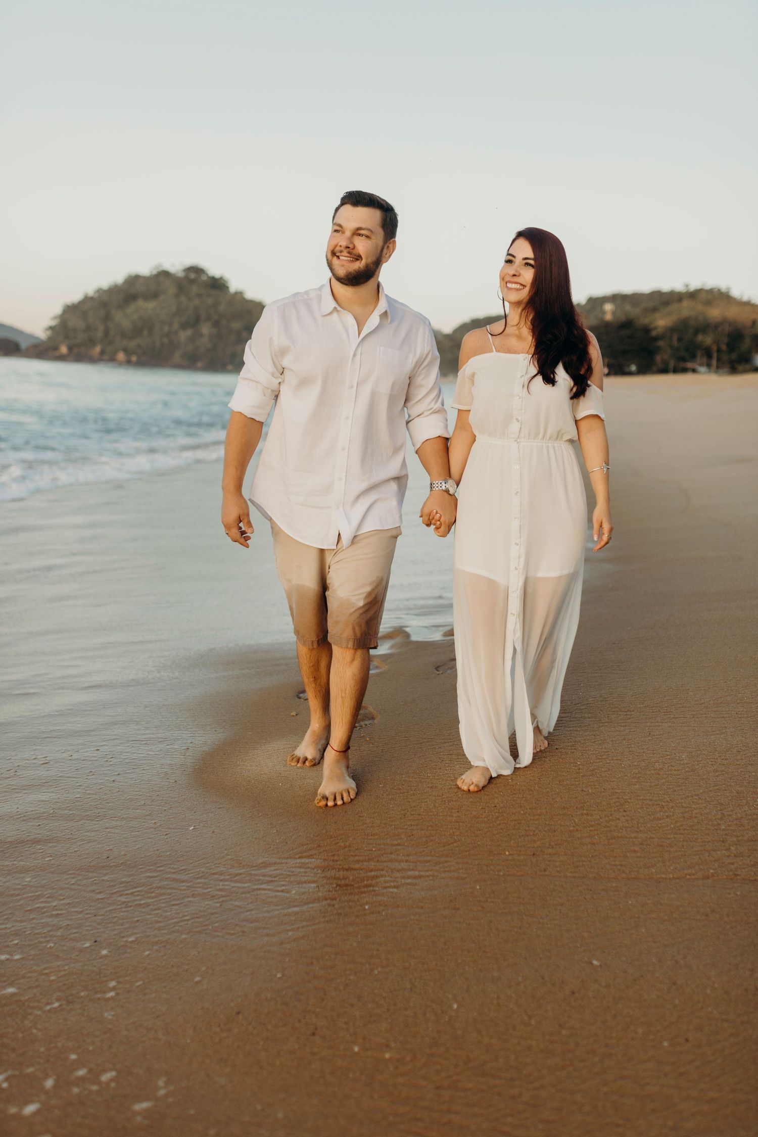 Couple in matching white outfits walking barefoot on beach during golden hour.