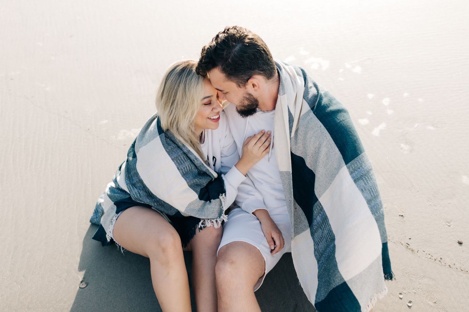 Couple sharing tender moment on beach while wrapped in cozy striped blanket.