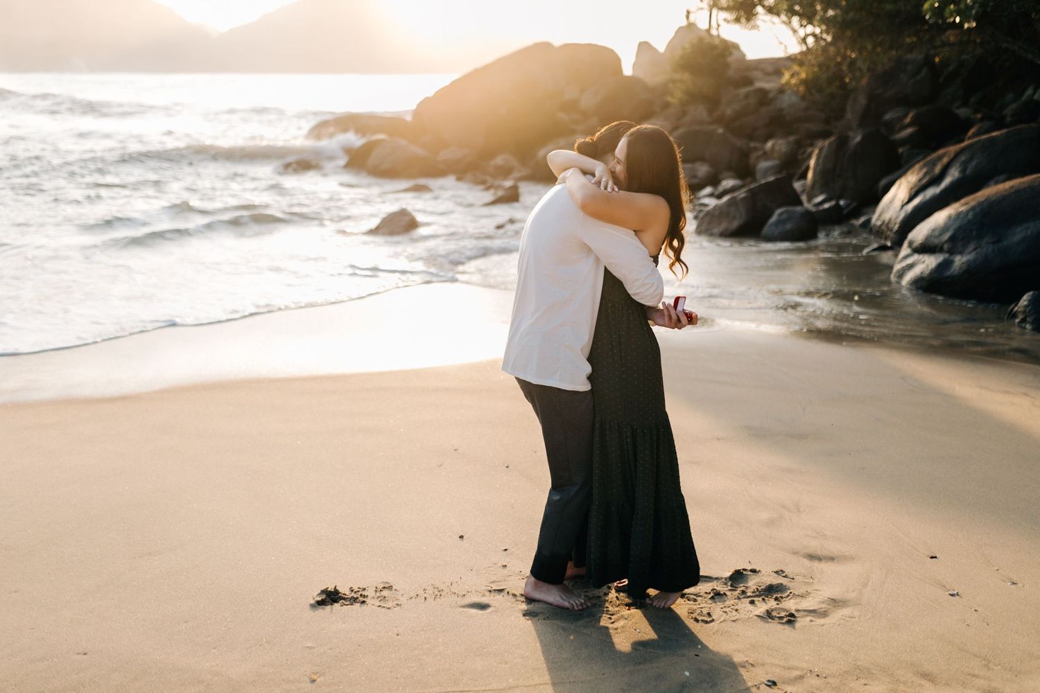 Joyful embrace after beach proposal with dramatic coastal scenery at sunset.