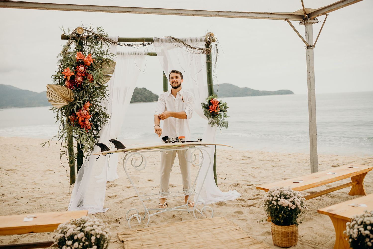 Beachside wedding ceremony setup with wooden benches and tropical floral arrangements under a white canopy.