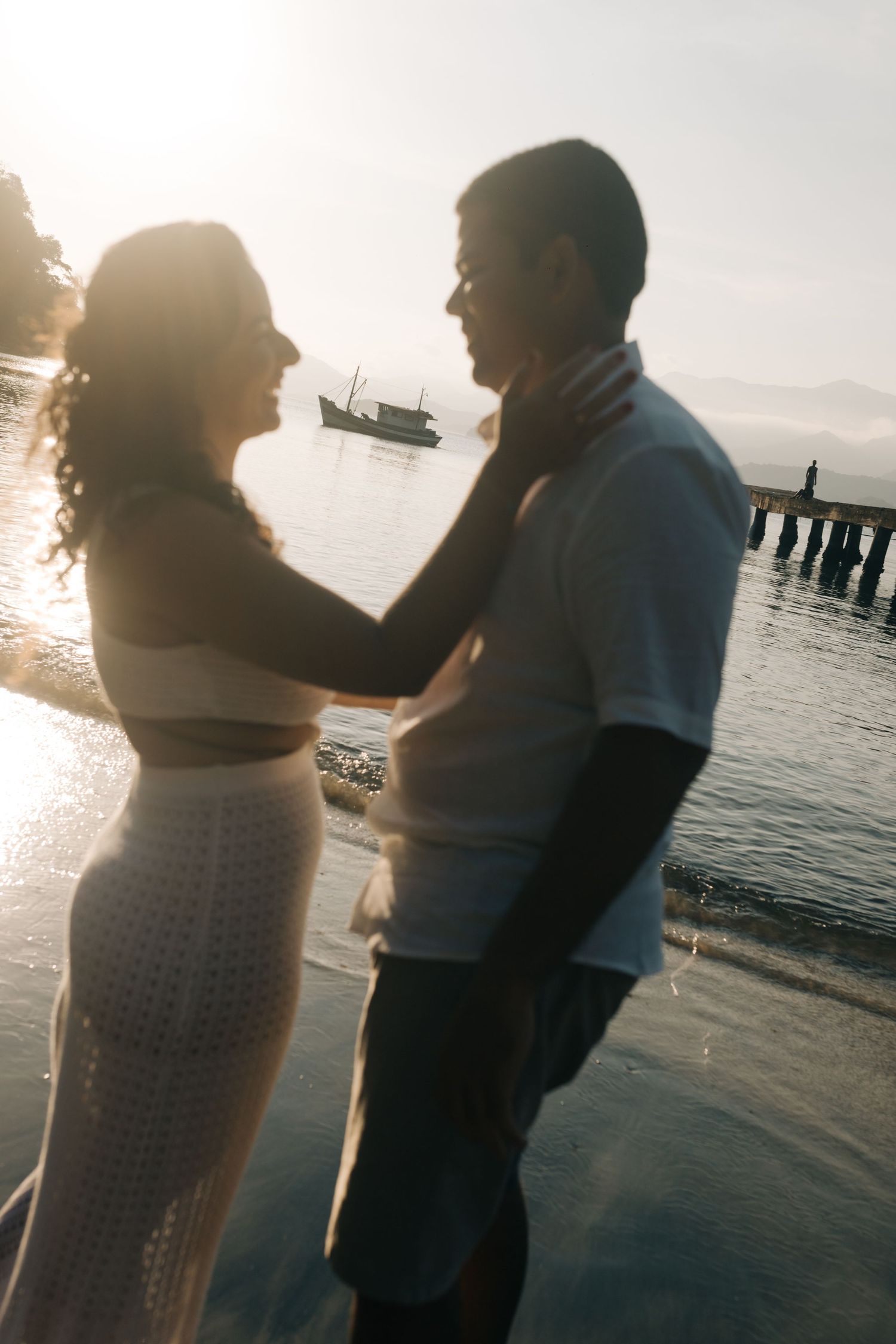 Couple sharing tender moment on beach at sunset with ocean and pier in background.