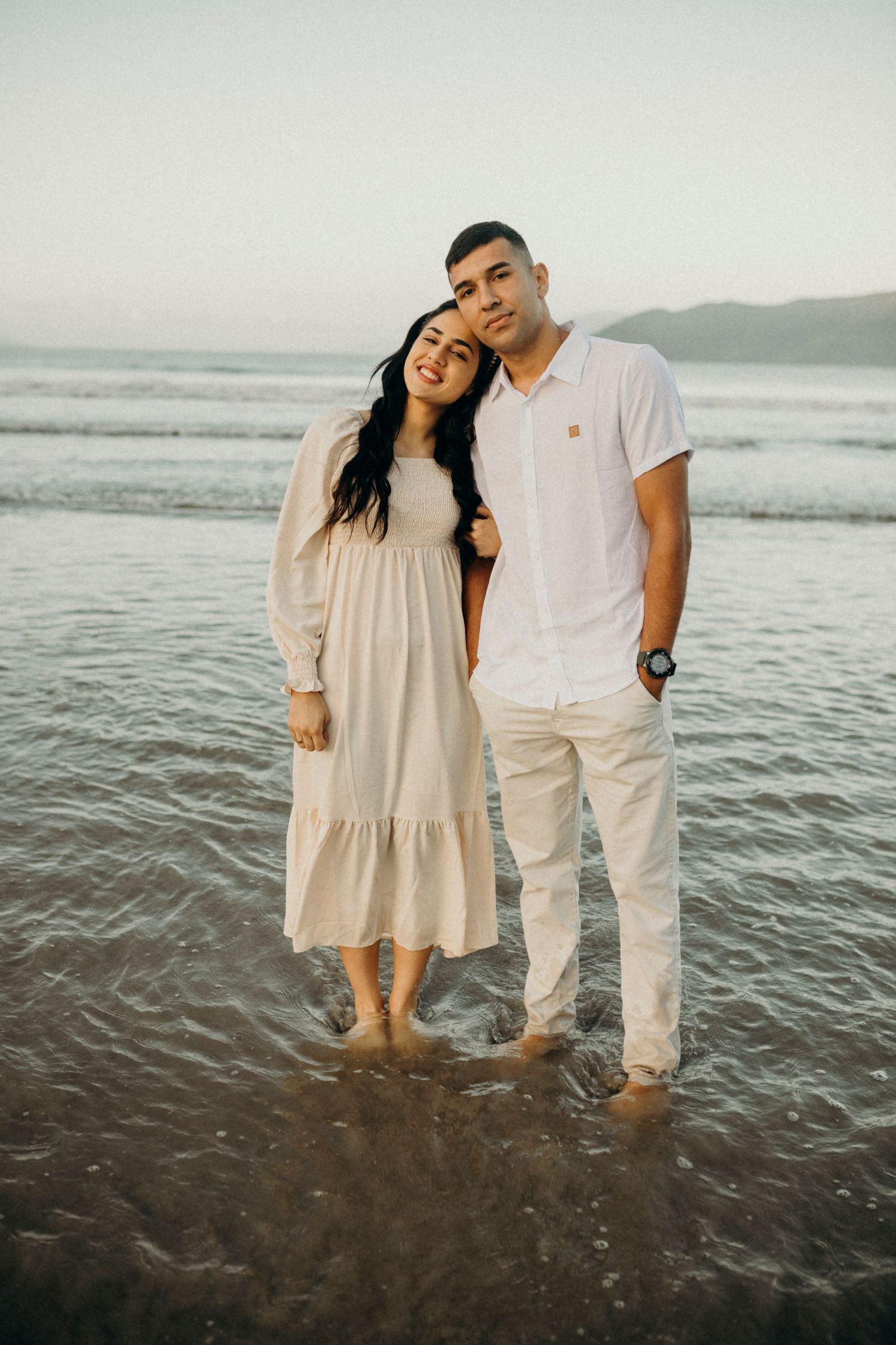 A couple dressed in white stands together in shallow ocean water during a beach photoshoot.