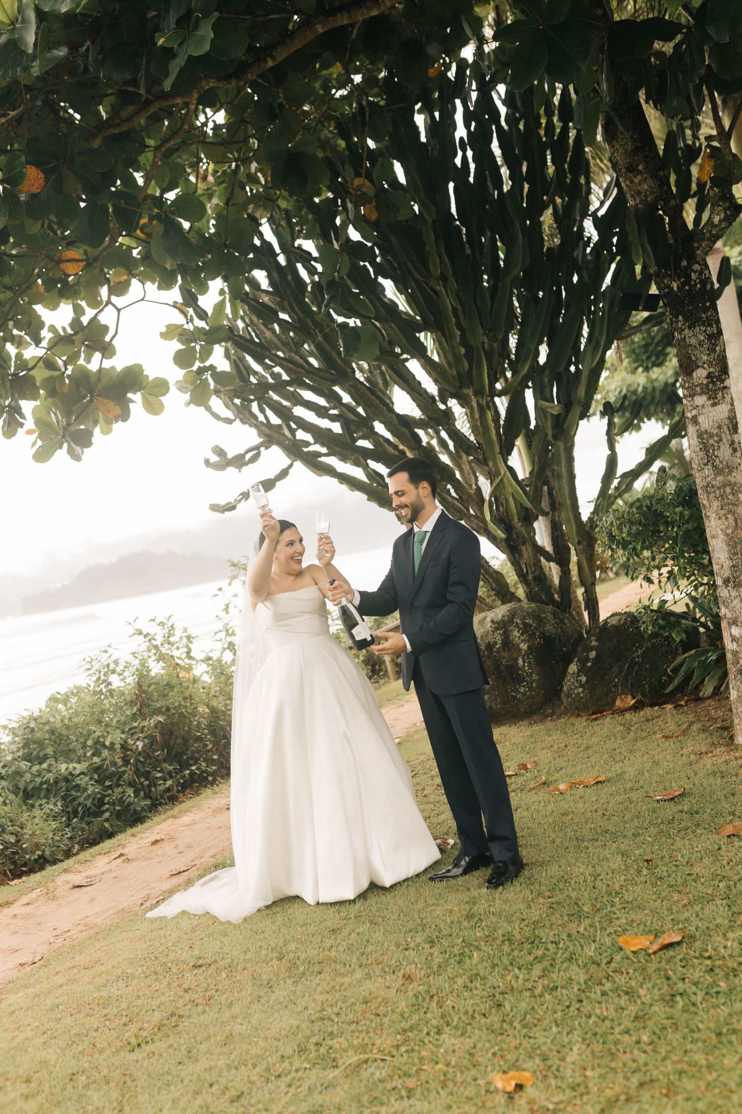 Happy couple celebrates with champagne under a tree during their coastal wedding.