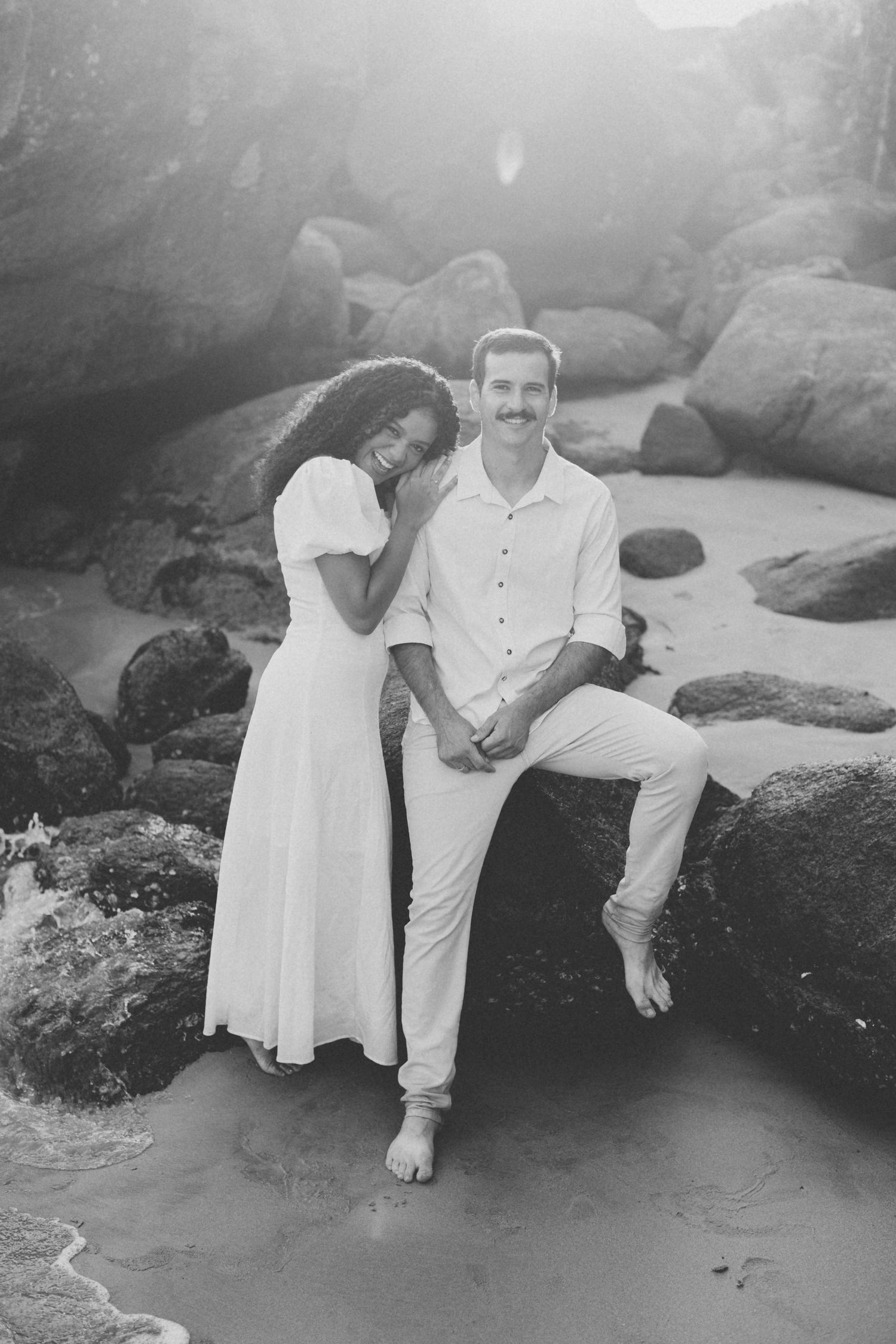 A romantic black and white photo of a couple posing on coastal rocks near the ocean.