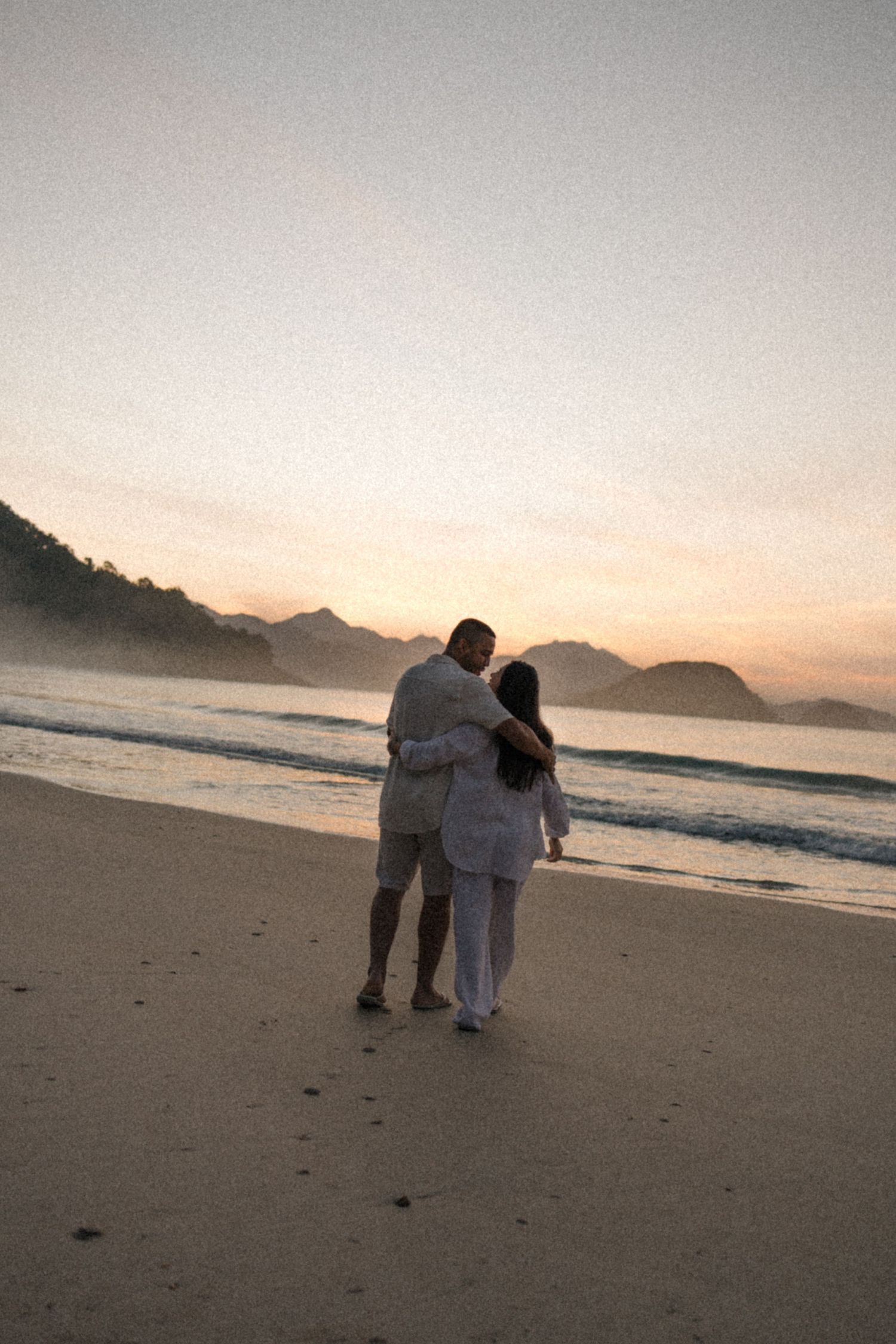 Silhouetted couple embracing on beach shore with mountainous coastline at dusk.