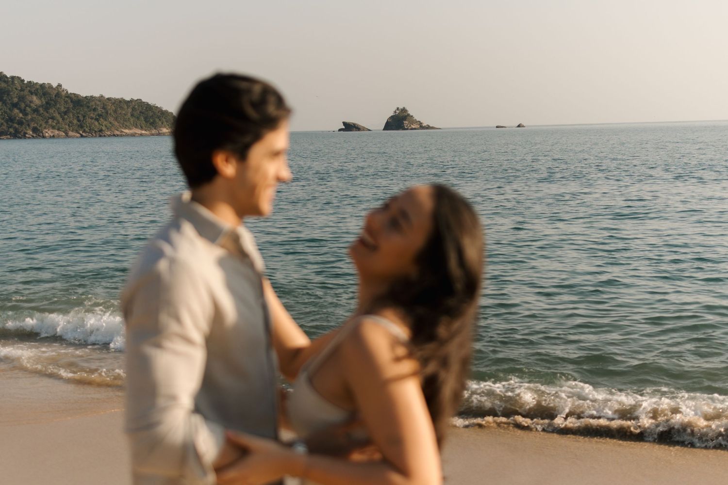 A couple shares an intimate moment together on a scenic beach with rolling waves and mountains in the background.