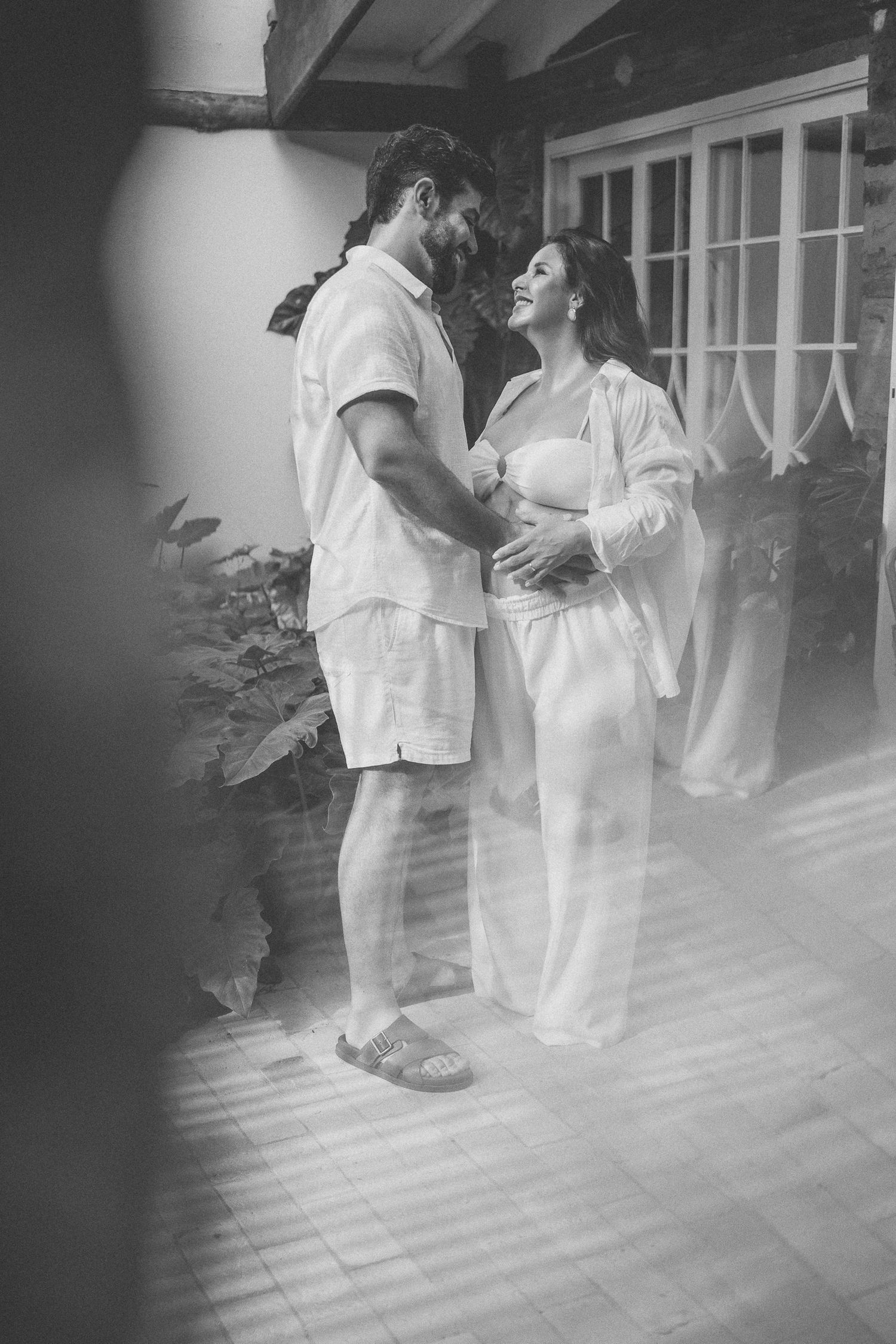 A romantic black and white photograph of a couple in white clothing embracing on a porch with French doors in the background.