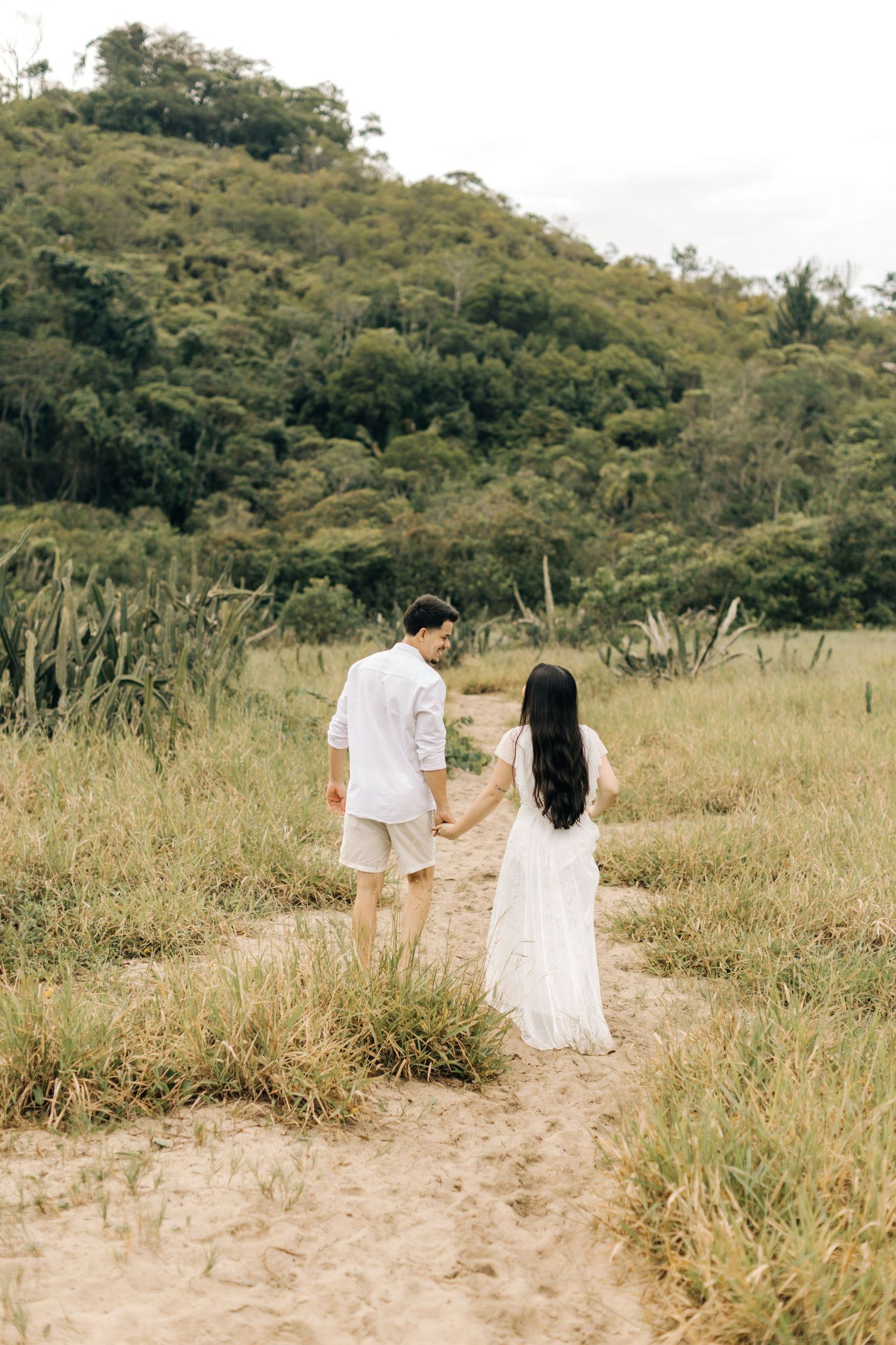A couple in white outfits holds hands while walking through a grassy field with hills in the background.