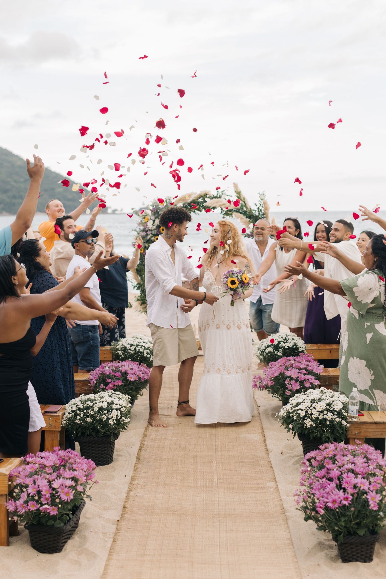 Wedding guests throw red rose petals as newlyweds walk down a burlap aisle runner on the beach.