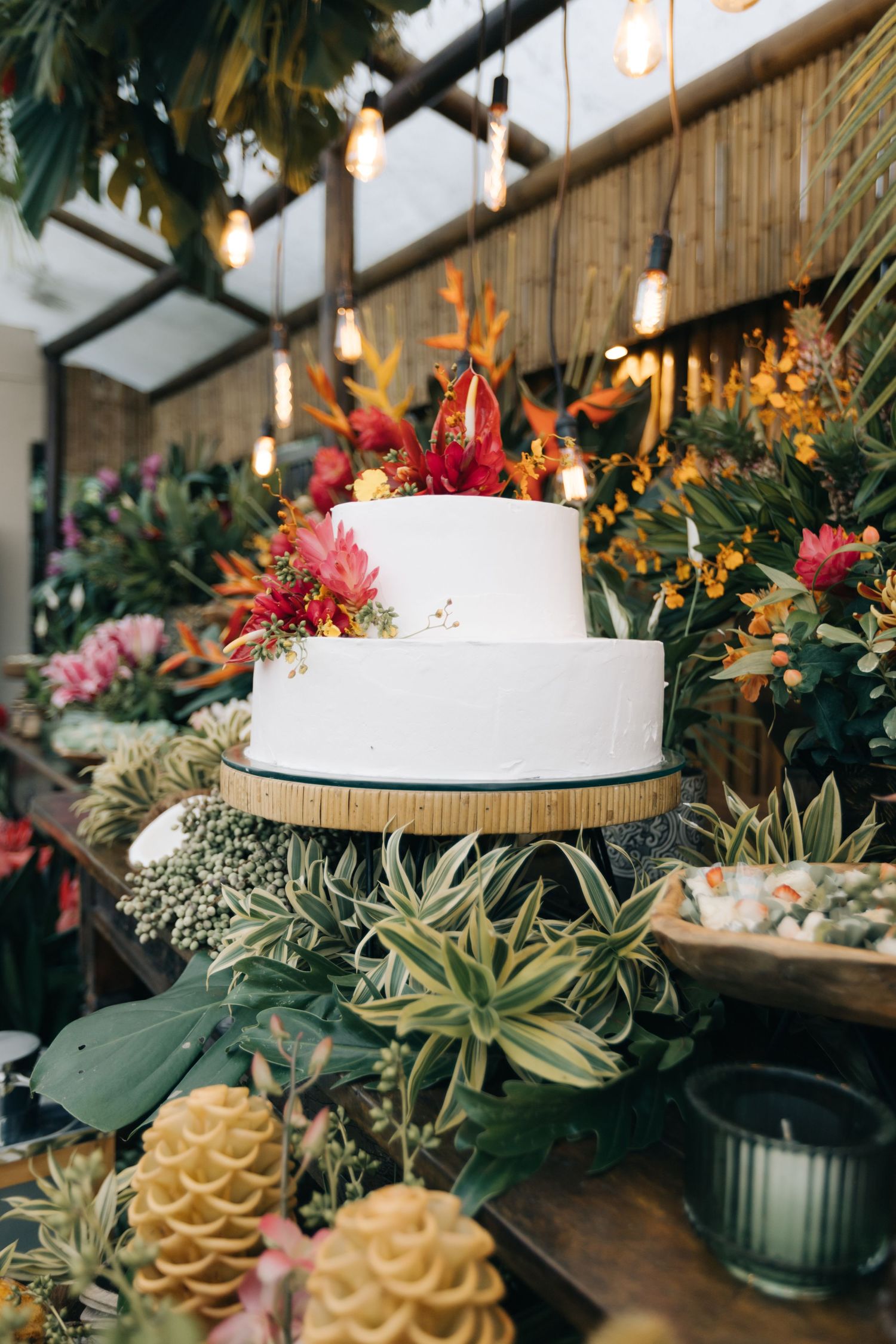 White wedding cake decorated with tropical flowers and succulents displayed on a wooden table with greenery.