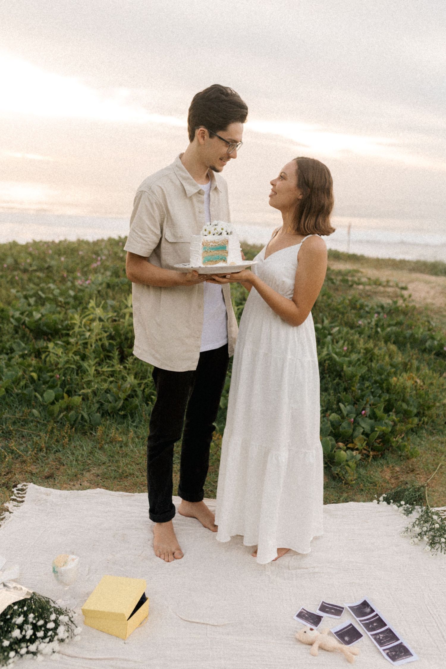 Couple enjoying cake and cards together on a white blanket in a grassy outdoor setting at sunset.