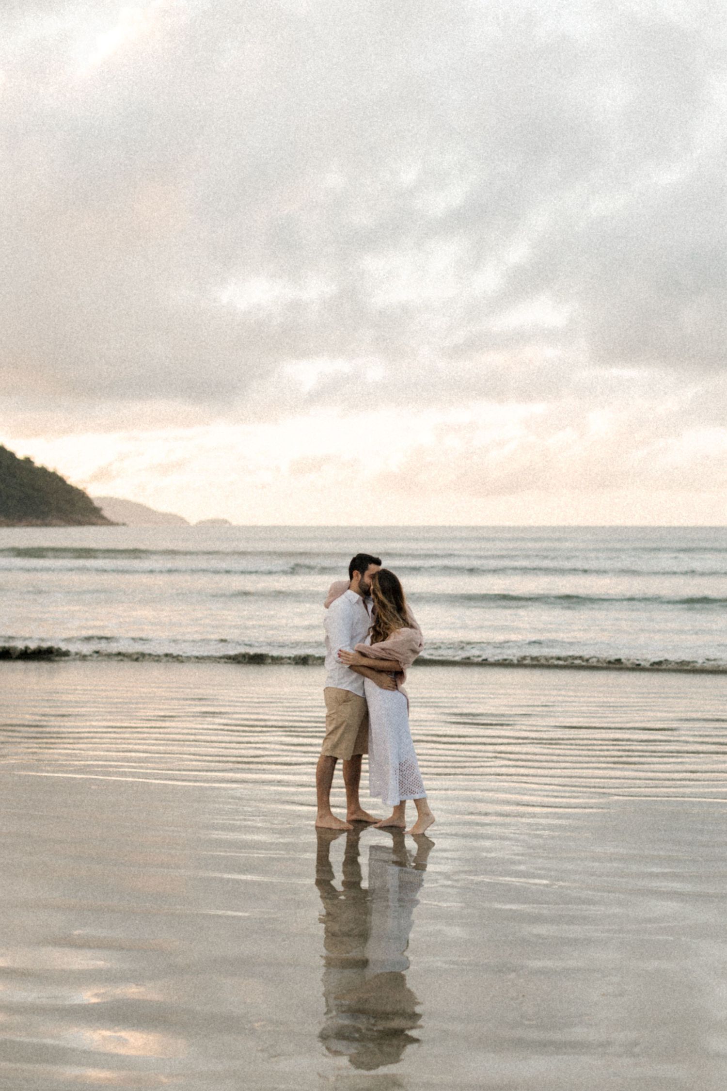 Couple stands together on reflective wet sand at sunset with coastal mountains in background.