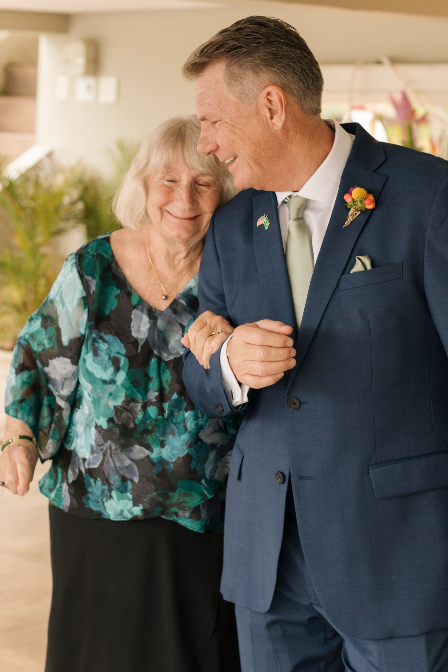 Two people share an emotional moment at a wedding, one wearing a navy suit and the other in a teal patterned top.