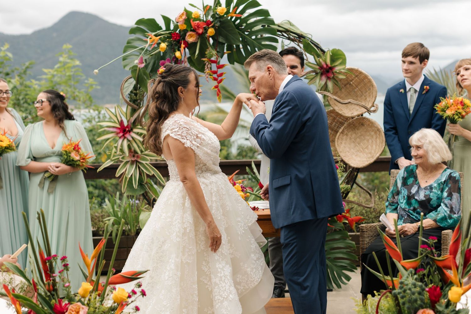 Outdoor wedding ceremony with mountain backdrop, tropical flowers, and guests in formal attire.