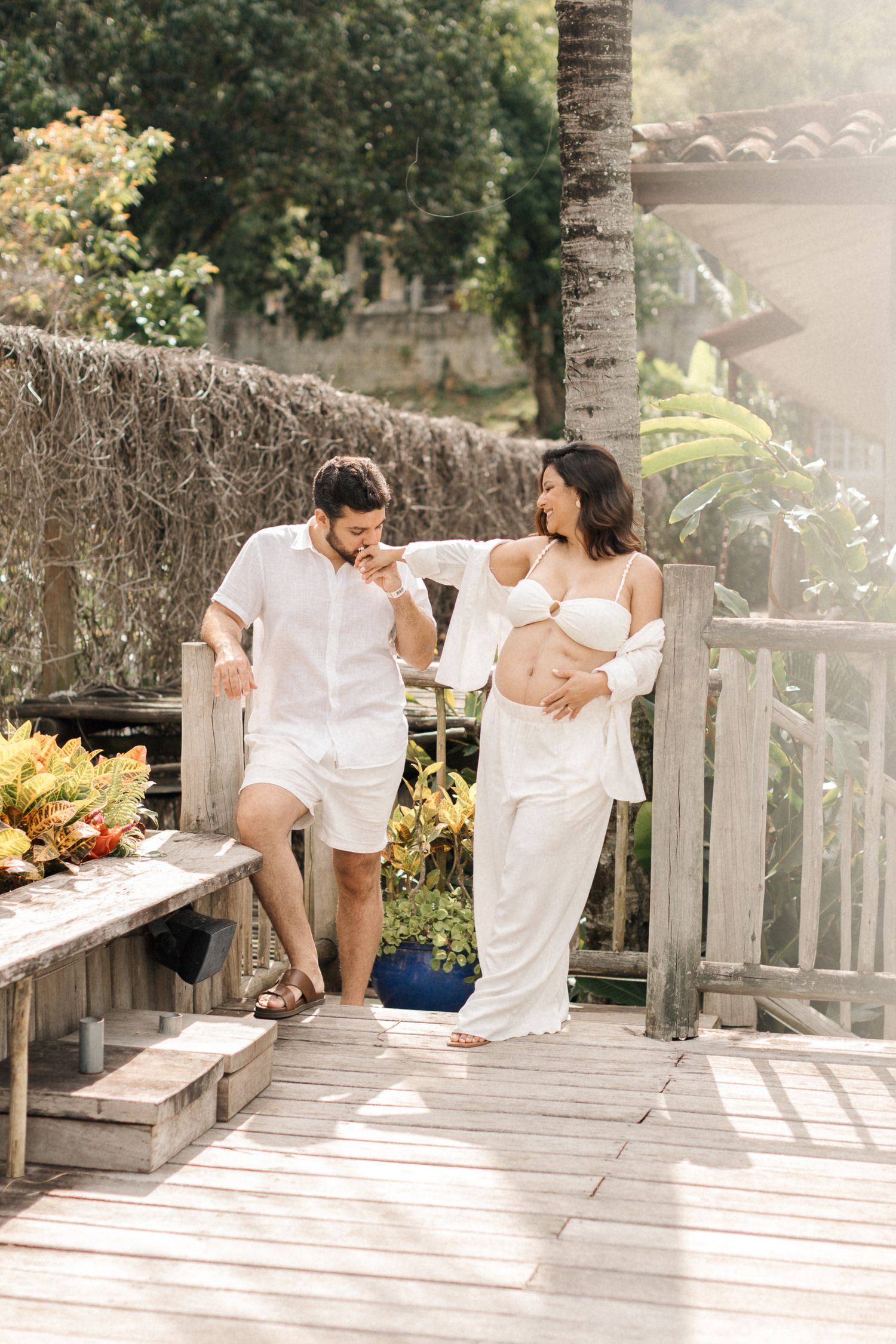 A couple in white clothing sits together on a wooden deck surrounded by lush greenery and palm trees on a sunny day.