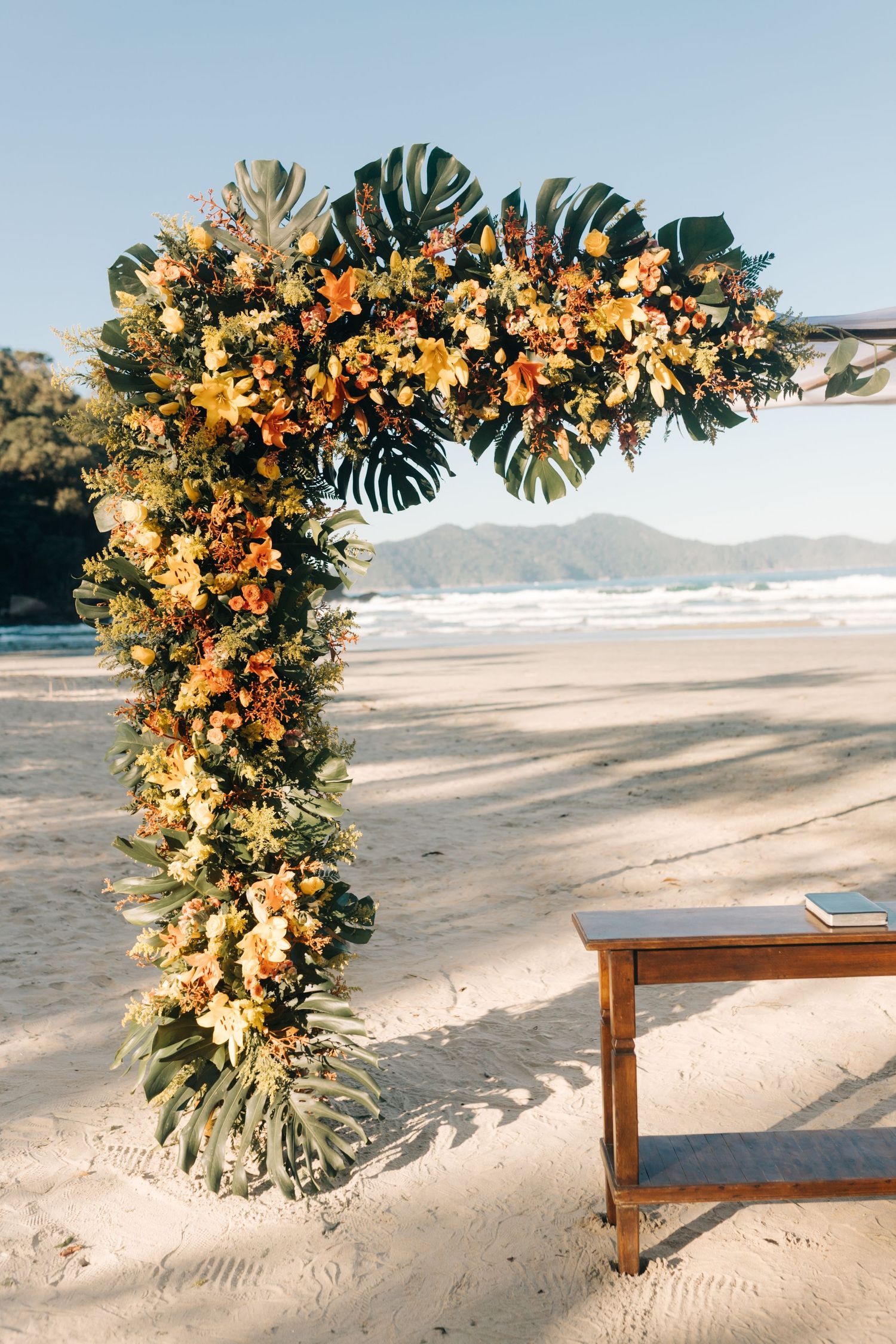 Close-up of wedding arch decorated with monstera leaves and orange flowers on sandy beach.