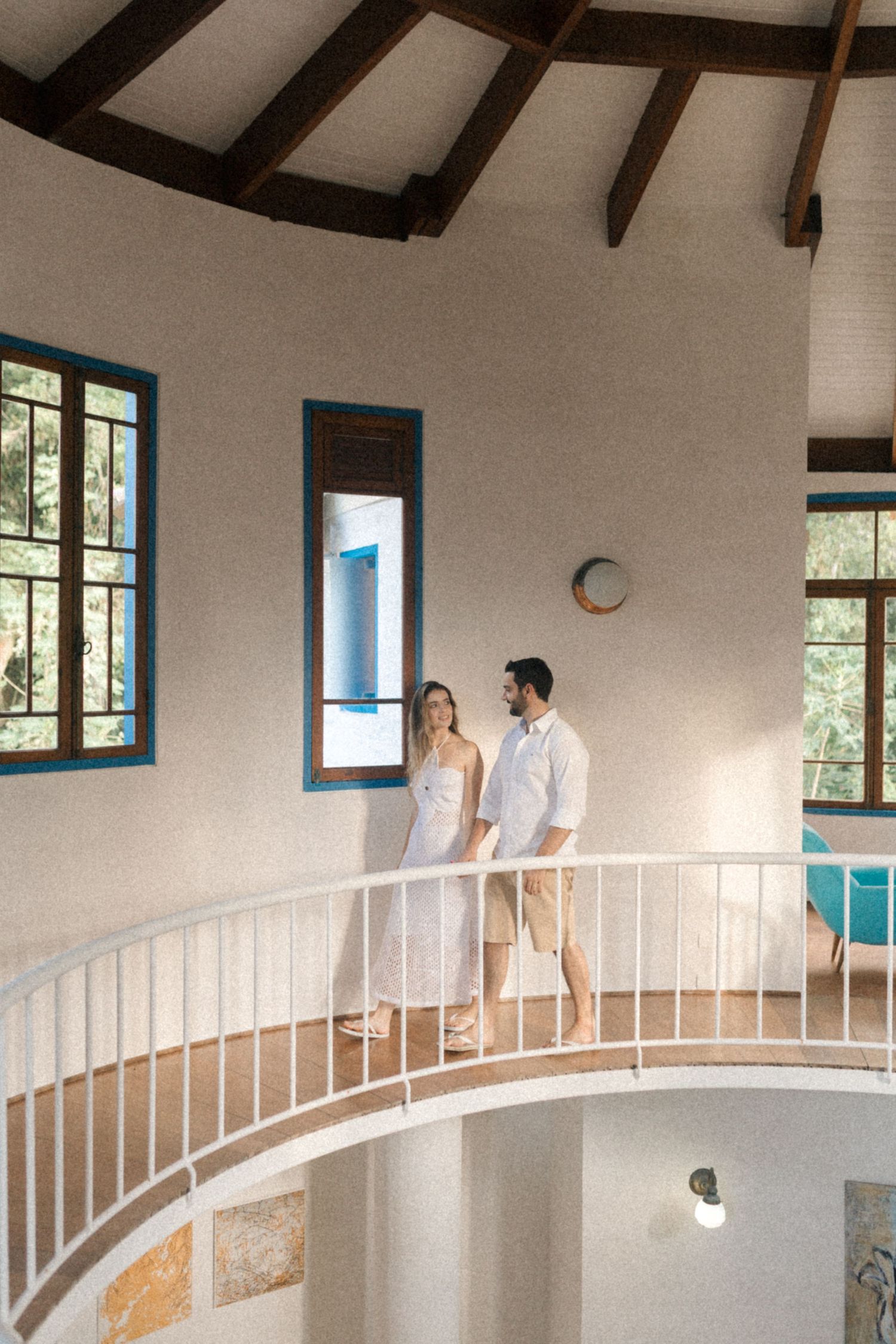 A couple stands together on a white railed balcony inside a circular room with wooden beams.