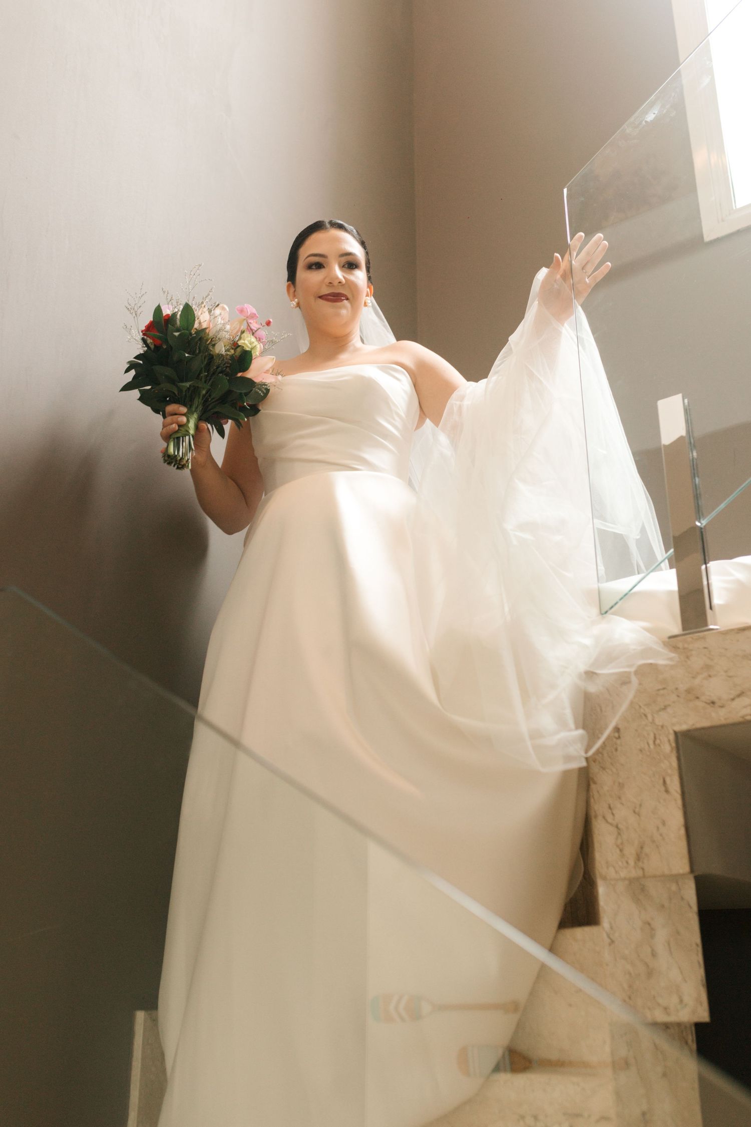 A bride in a white strapless gown poses with a pink bouquet on an elegant staircase.